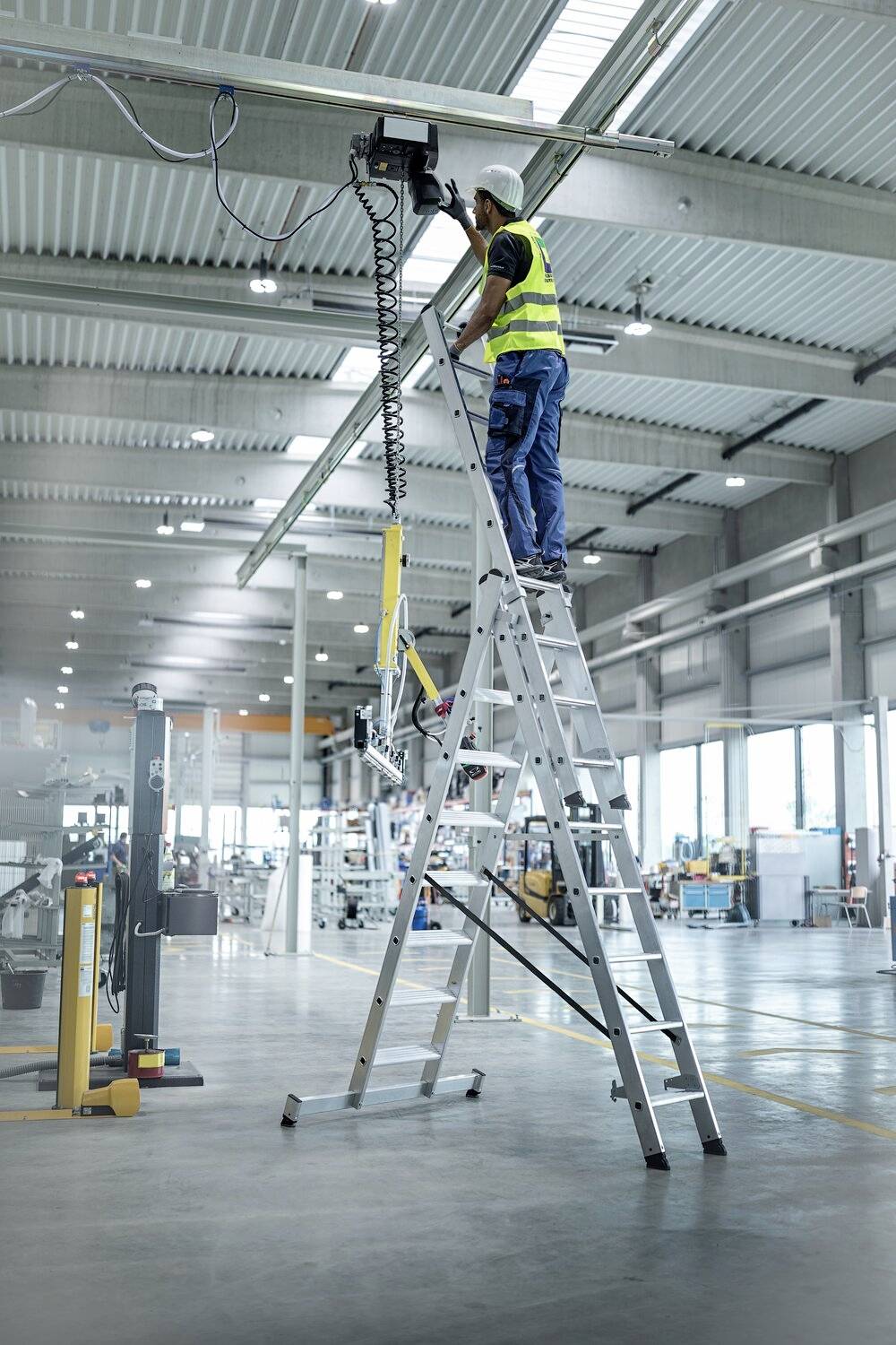 Eine Person mit Warnweste steht auf einer Leiter in einem großen, hellen Industriegebäude und arbeitet an Kabeln an der Decke.
