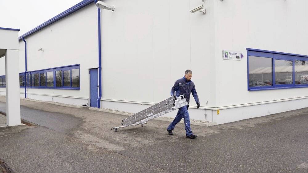 Ein Mann trägt eine lange Aluminiumleiter auf einem Industriegelände entlang eines weißen Gebäudes mit blauen Fenstern und Türen.