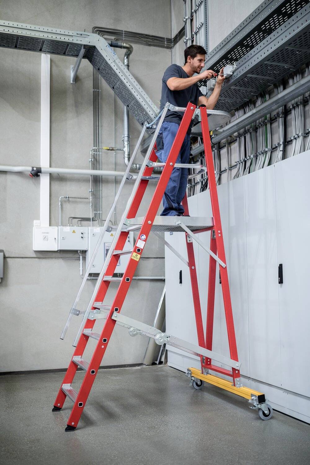 Ein Mann auf einer roten Klappleiter überprüft technische Kabel in einem Industriegebäude. Im Hintergrund sind Leitungen und Schaltschränke sichtbar.