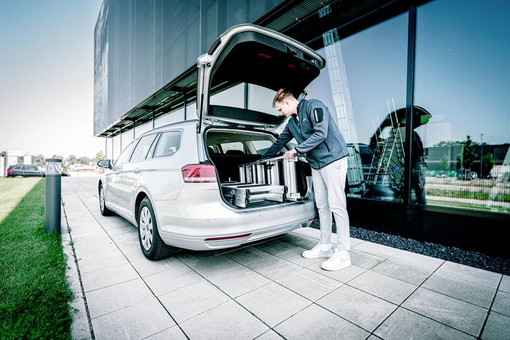 Ein Mann lädt Koffer in den offenen Kofferraum eines silbernen Autos vor einem modernen Gebäude.