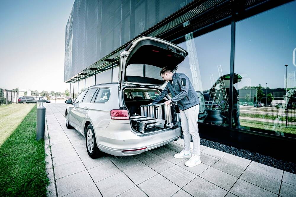 Eine Person lädt Gegenstände in den Kofferraum eines silbernen Kombis vor einem modernen Gebäude mit Glasfassade.