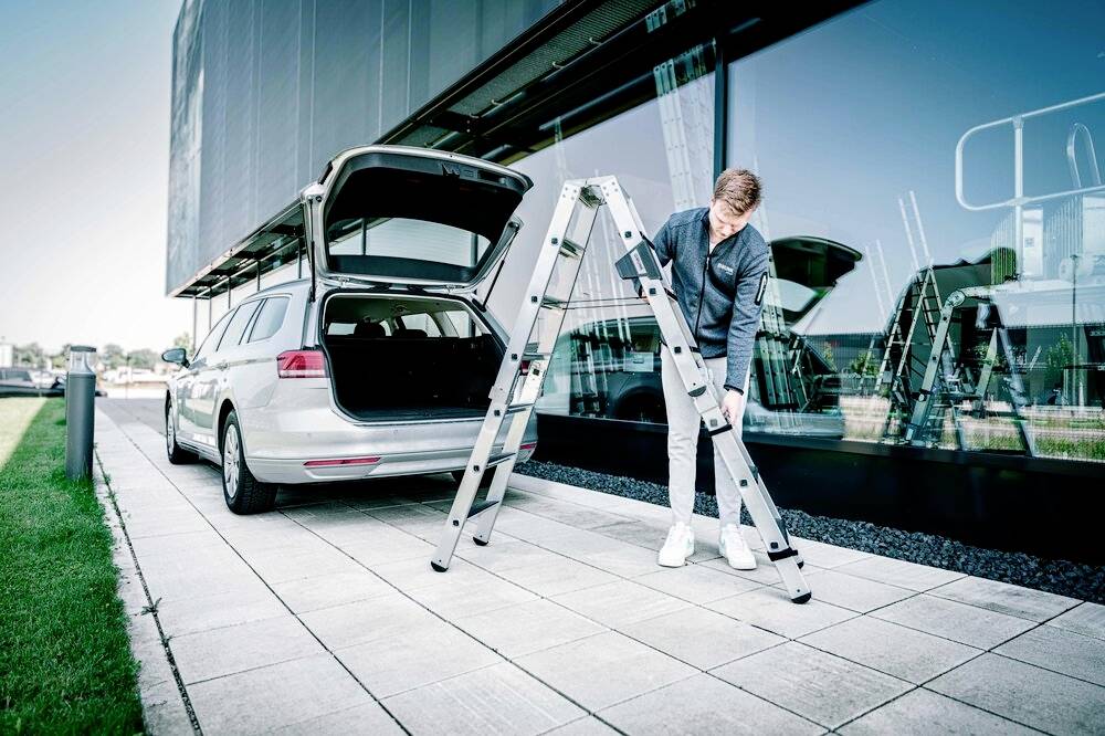 Eine Person stellt eine klappbare Leiter neben einem geparkten Kombi auf. Die Heckklappe des Autos ist geöffnet.