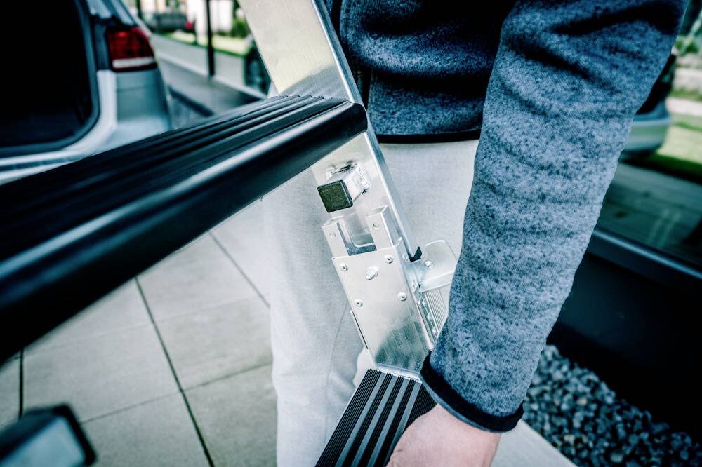 Eine Person trägt eine zusammengeklappte Leiter. Im Hintergrund ist ein Auto zu sehen.
