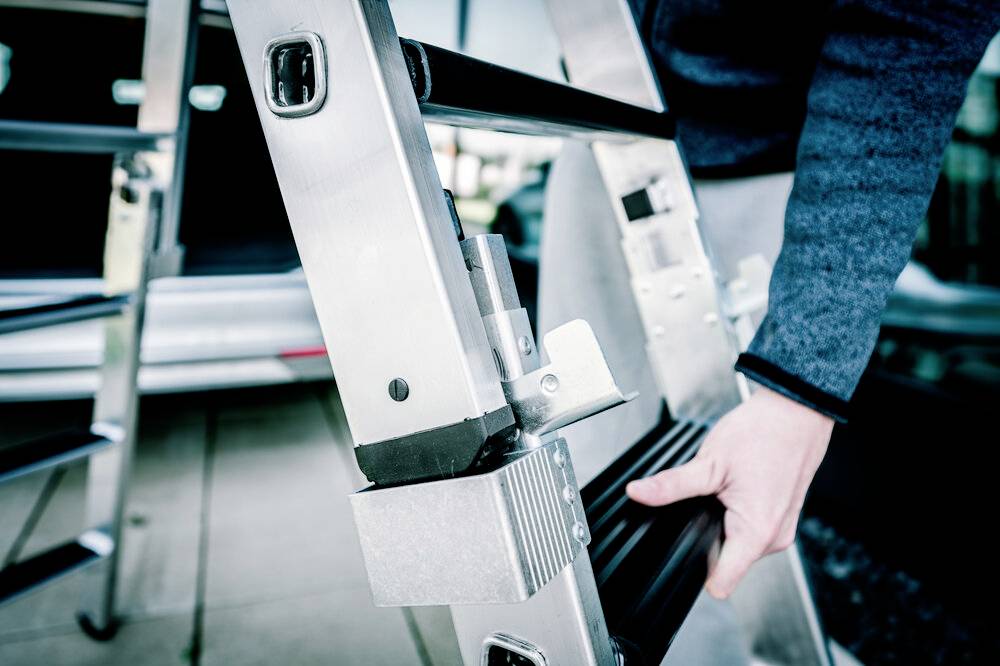 Eine Person hält eine Metallleiter; der Fokus liegt auf den Händen und der Leiter. Im Hintergrund sind unscharfe Autos sichtbar.