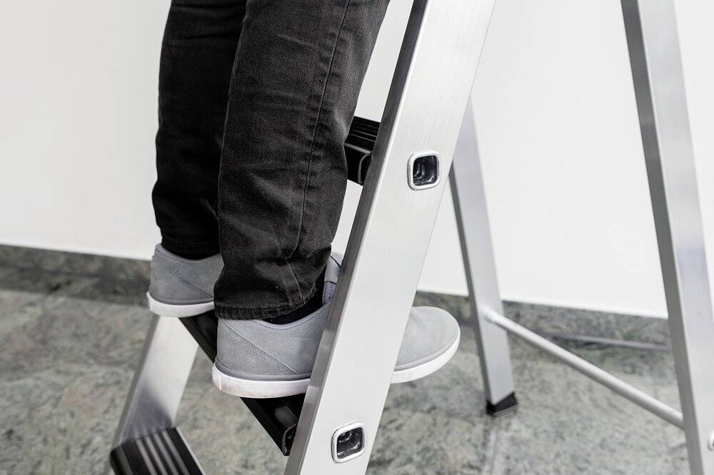 Eine Person in Jeans und grauen Schuhen steht auf einer Aluminiumleiter, bereit zum Aufstieg. Der Boden ist aus Marmor.