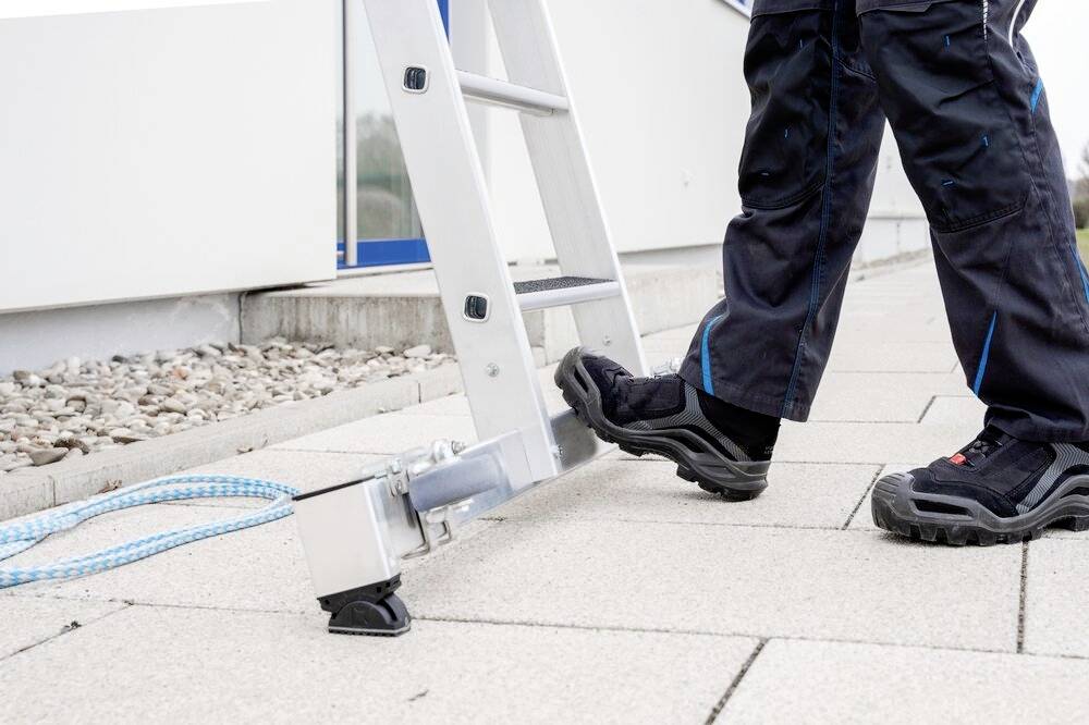 Eine Person in Arbeitshose und Sicherheitsstiefeln steht auf einer Leiter, die auf einem gepflasterten Boden mit einem Seil gesichert ist.