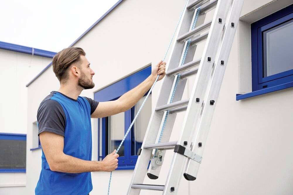 Ein Mann in blauem T-Shirt hält das Seil einer angelehnten Aluminiumleiter an einer Hauswand mit blauen Fenstern.