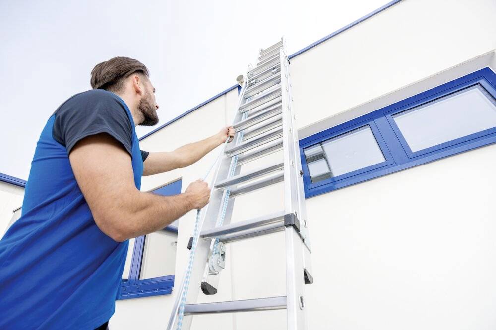 Eine Person klettert eine Leiter hinauf, die an einer Hauswand mit blauen Fenstern steht.