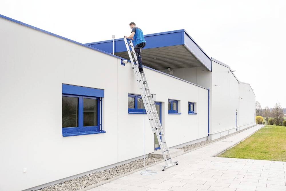 Person auf Leiter repariert Dach eines weißen Gebäudes mit blauen Fenstern.