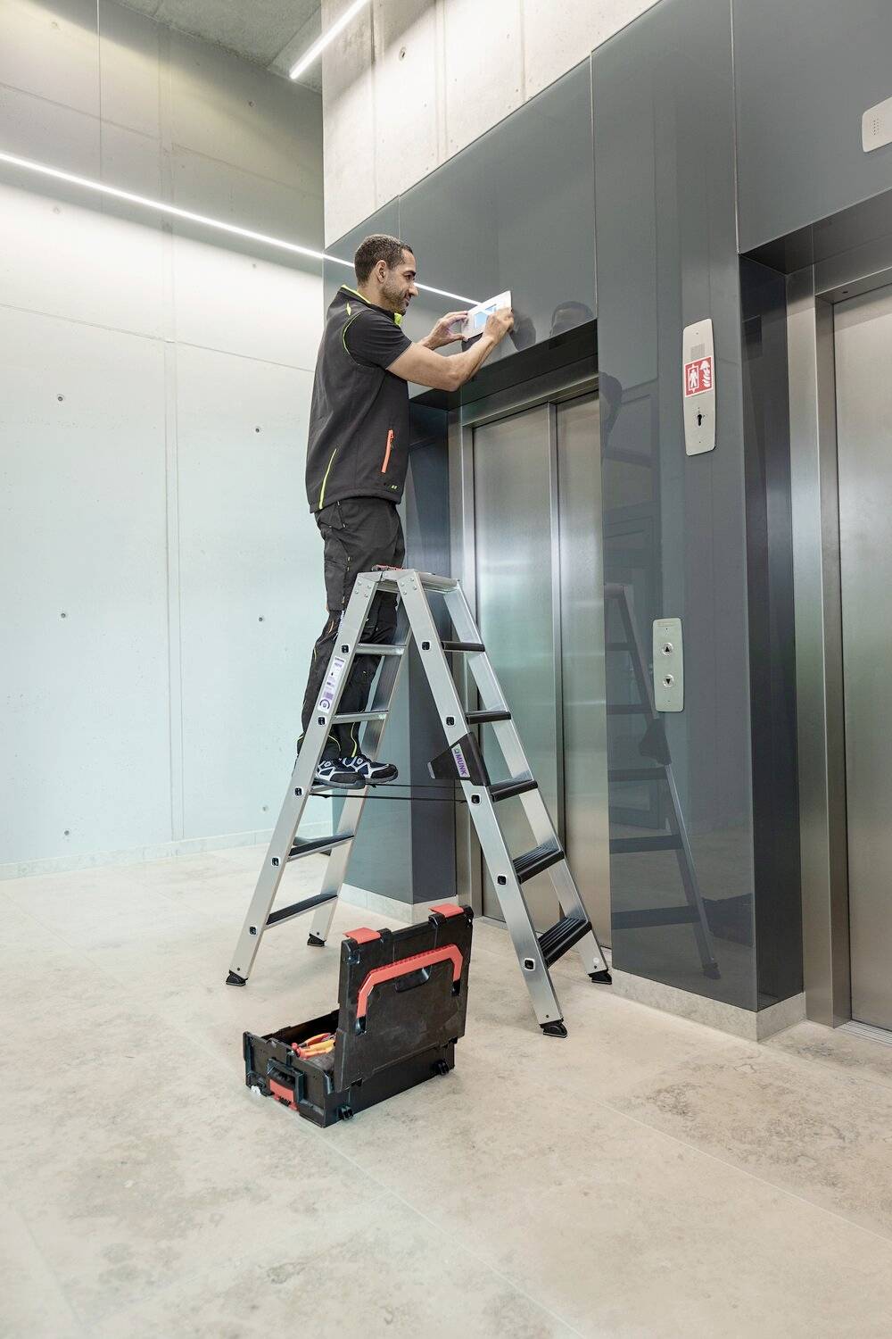 Ein Techniker steht auf einer Leiter und arbeitet oberhalb eines Aufzugs in einem modernen Gebäude. Neben dem Aufzug steht ein Werkzeugkoffer.