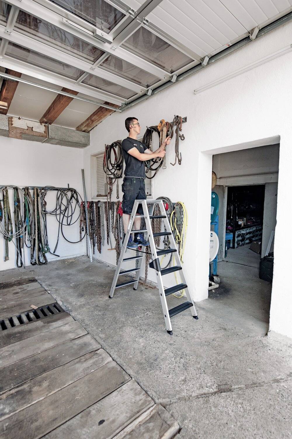 Eine Person steht auf einer Leiter in einer Garage und hängt Seile auf. An der Wand sind bereits mehrere Seile und Kabel befestigt.