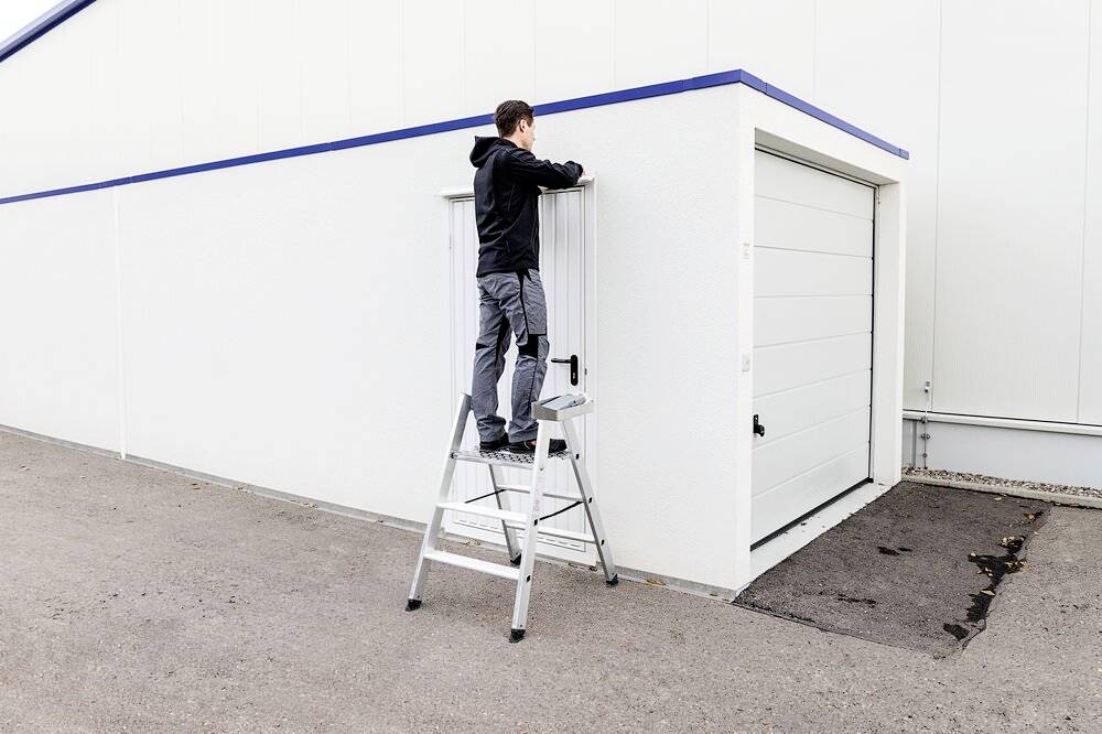 Eine Person steht auf einer Leiter und repariert oder installiert einen weißen Garagenanbau an einem Gebäude.