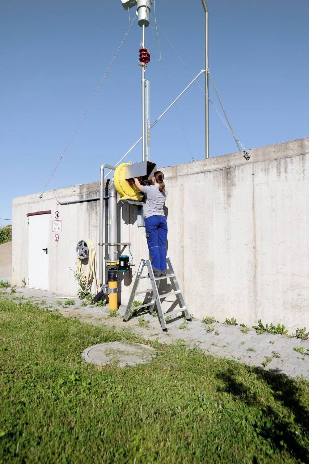 Person steht auf Leiter und arbeitet an gelbem Schutzschild auf einer Rohrleitung an einer Betonwand.