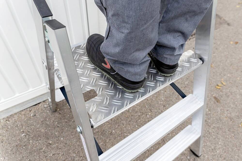 Person steht auf der obersten Stufe einer silbernen Metallleiter, die Schuhe tragen ein auffälliges Logo, Umfeld asphaltierter Boden.