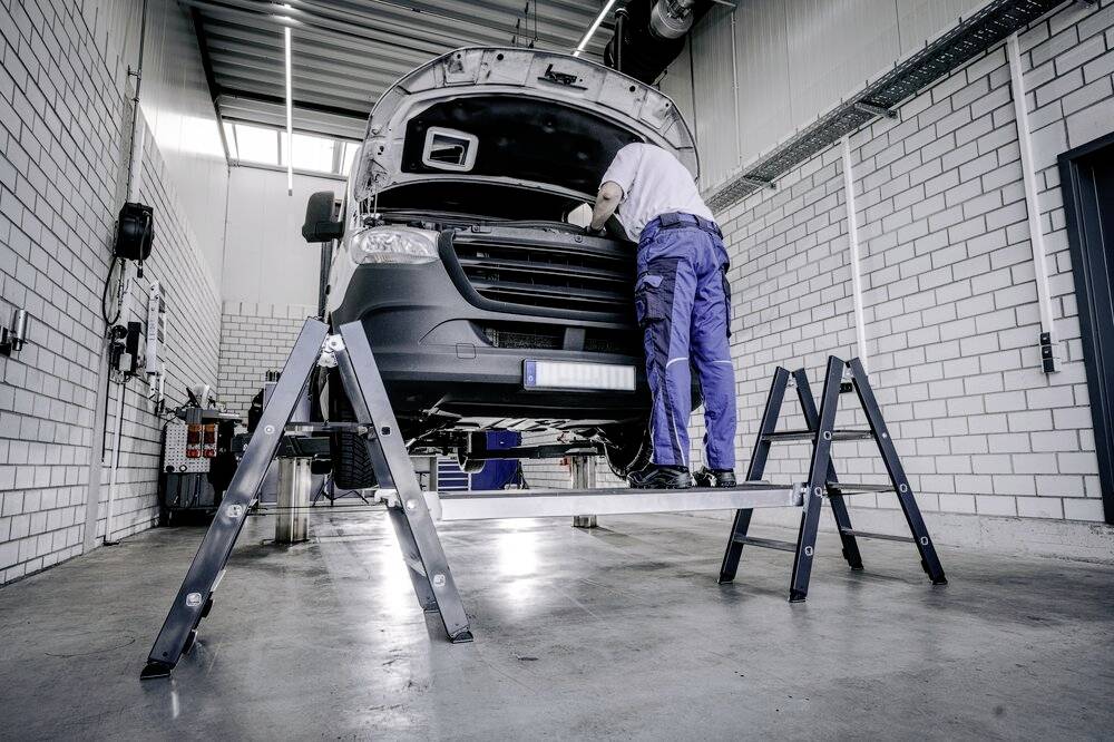 Mechaniker arbeitet in einer Werkstatt an einem gelifteten Fahrzeug. Er prüft den Motorraum. Umgebung zeigt industrielle Ausstattung.