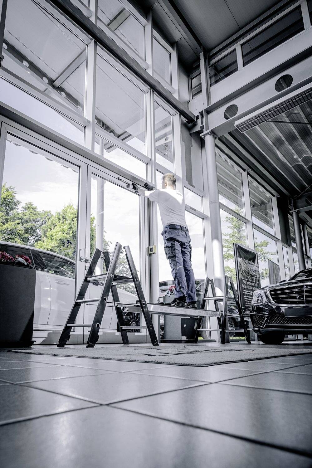 Person steht auf einer Leiter in einem Autohaus und reinigt ein hohes Fenster. Im Hintergrund stehen Autos, darunter ein schwarzer Wagen.