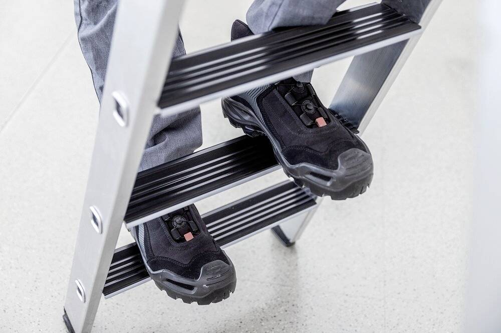 Eine Person in Arbeitskleidung steigt eine Aluminiumleiter hinauf, trägt schwarze Sicherheitsschuhe.