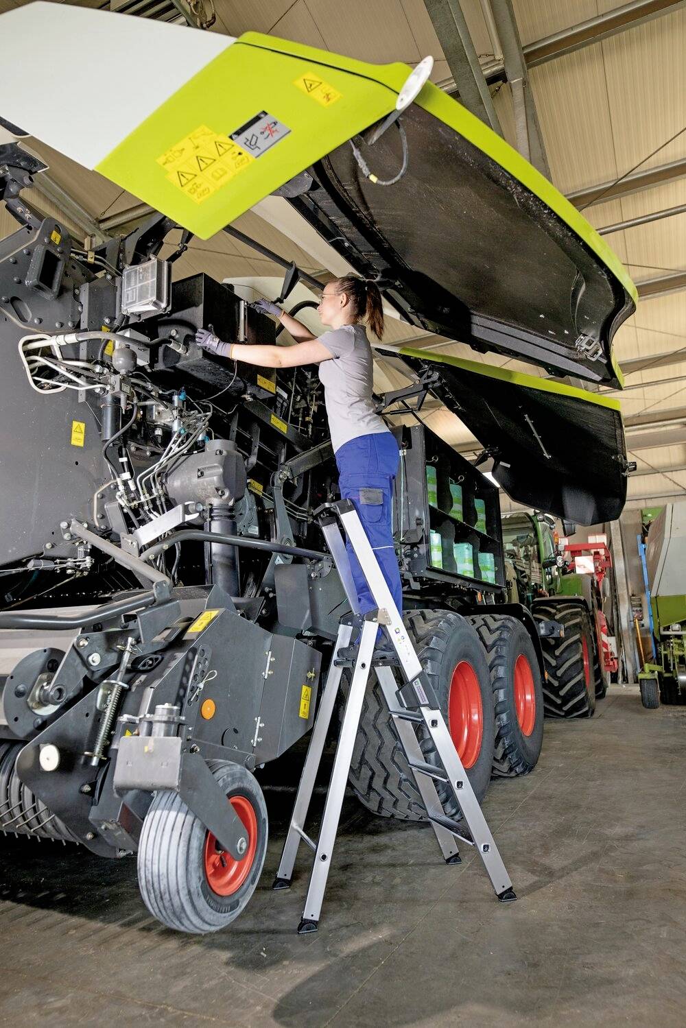 Eine Frau steht auf einer Leiter und arbeitet an einem großen landwirtschaftlichen Fahrzeug in einer Werkstatt.
