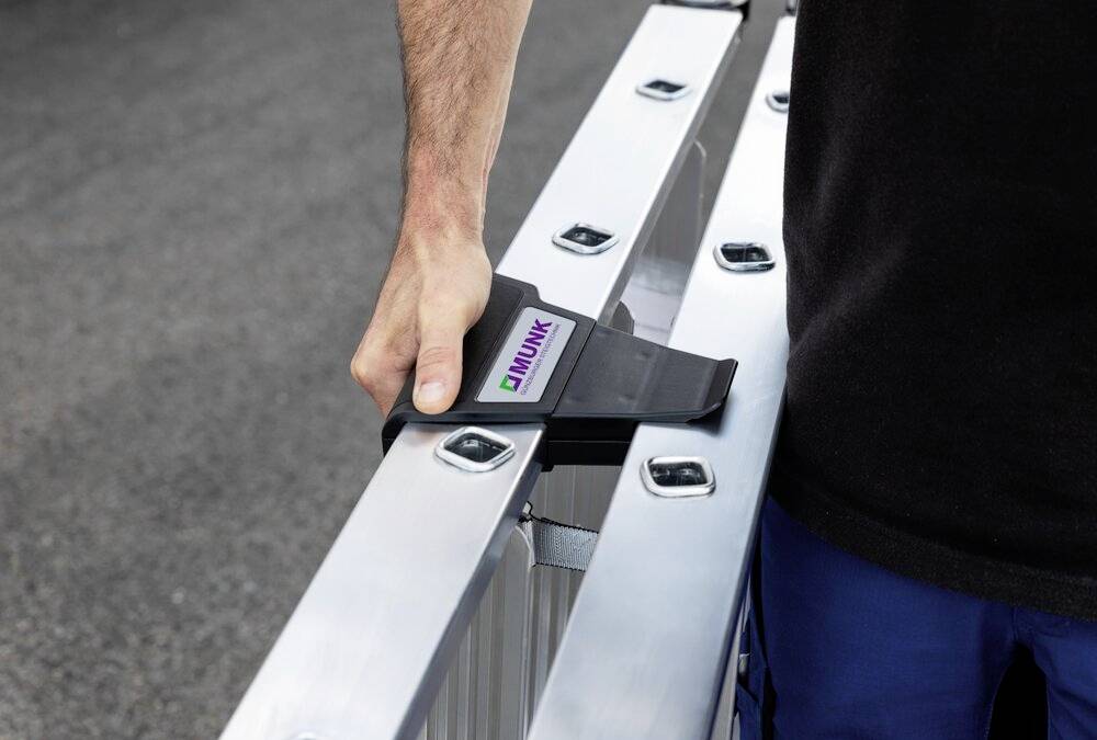 Eine Person hält eine Aluminiumleiter in der Hand. Auf der Leiter befindet sich ein Etikett mit dem Logo 'Munk'. Die Person trägt dunkle Kleidung.