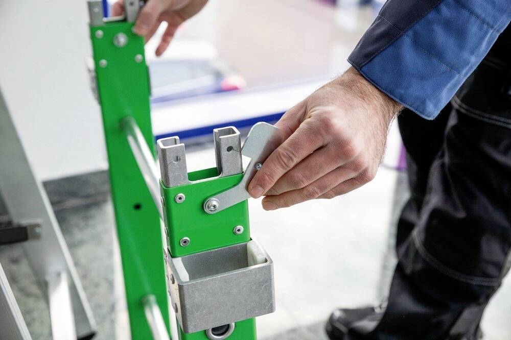 Eine Person justiert den Hebel einer grünen Treppe oder Plattform. Fokus auf den Mechanismus. Hintergrund unscharf.