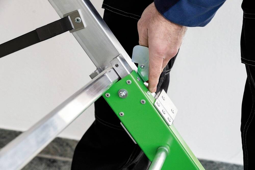 Ein Handwerker hält einen grünen Klappmechanismus einer Aluminiumleiter fest, zeigt die Handhabung und Sicherheit des Leiterverschlusses.