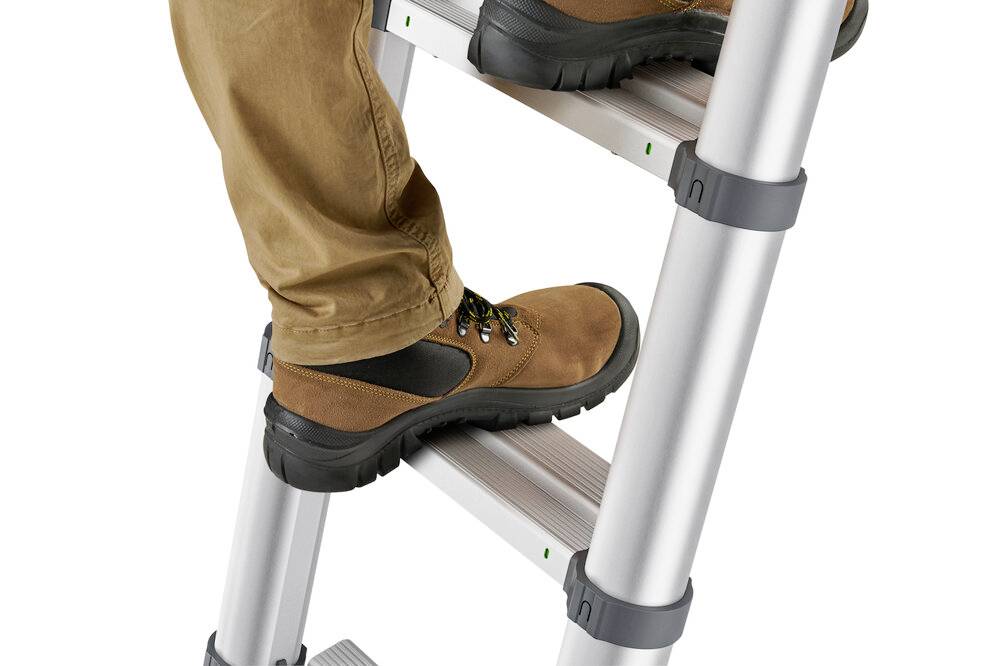 Eine Person steigt eine Aluminiumleiter hinauf, trägt braune Schuhe und beige Hosen. Fokus liegt auf den Füßen.