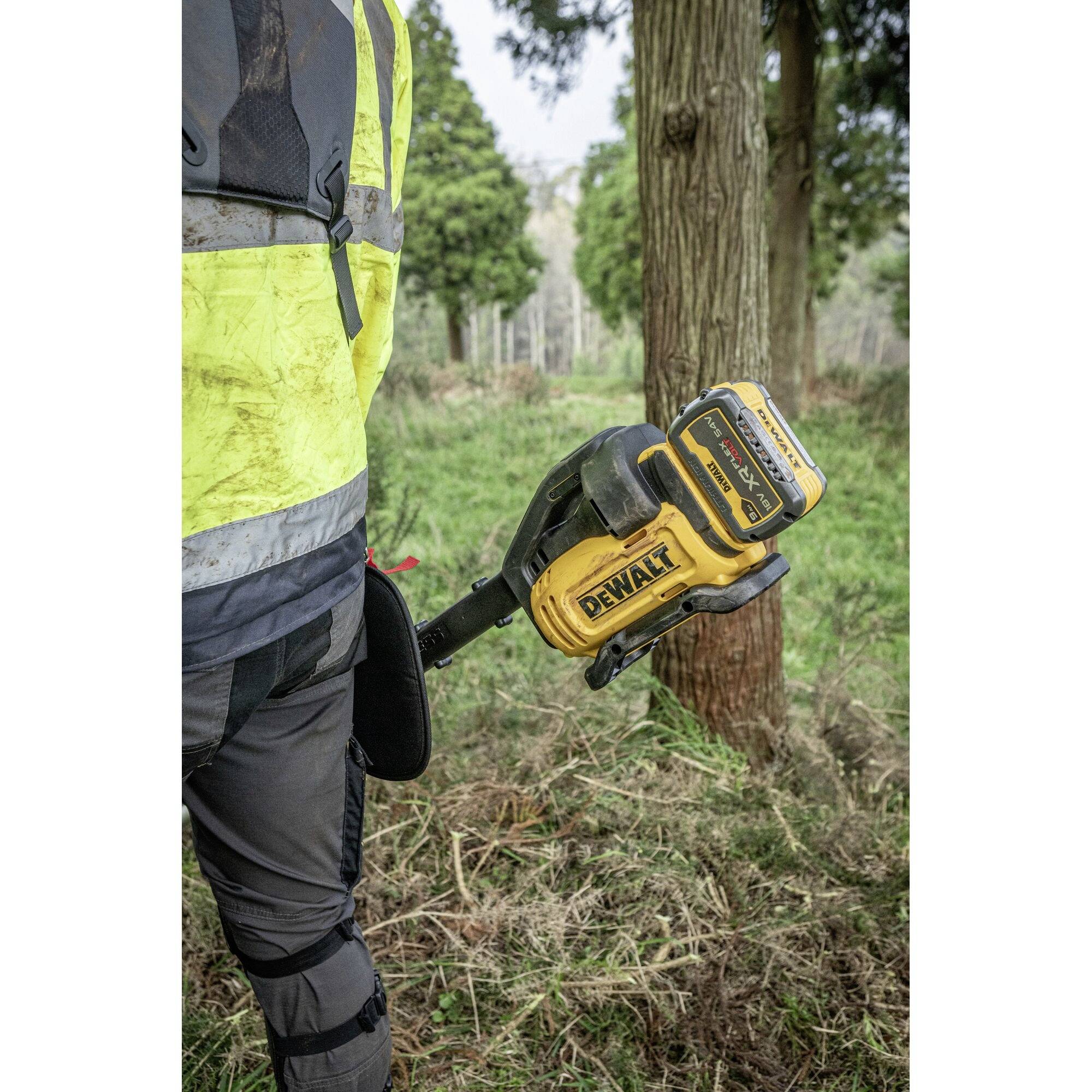 Eine Person in Arbeitskleidung hält ein gelbes batteriebetriebenes Werkzeug im Wald.
