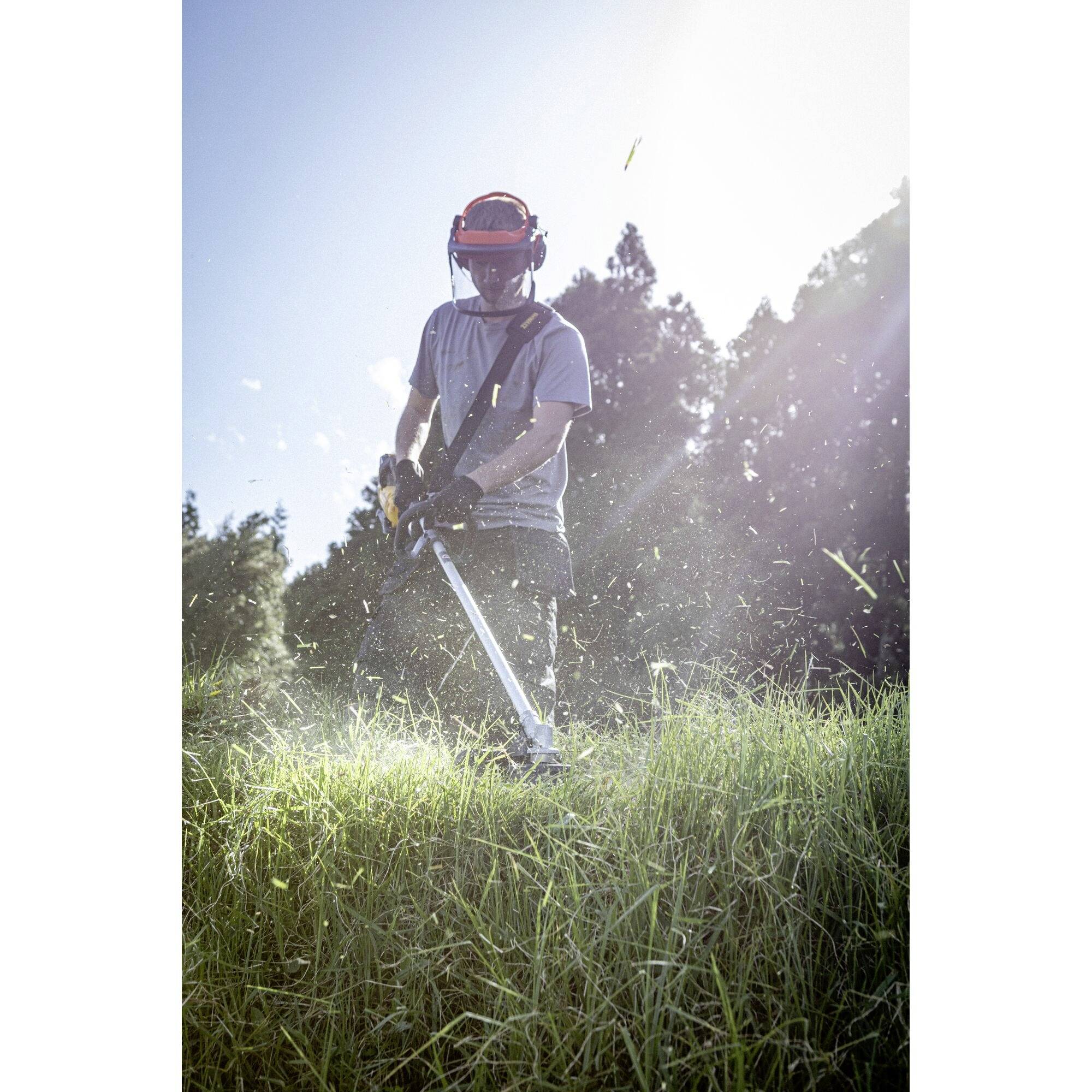 Eine Person mit Schutzkleidung mäht Gras mit einem Rasentrimmer in einem sonnigen Garten.