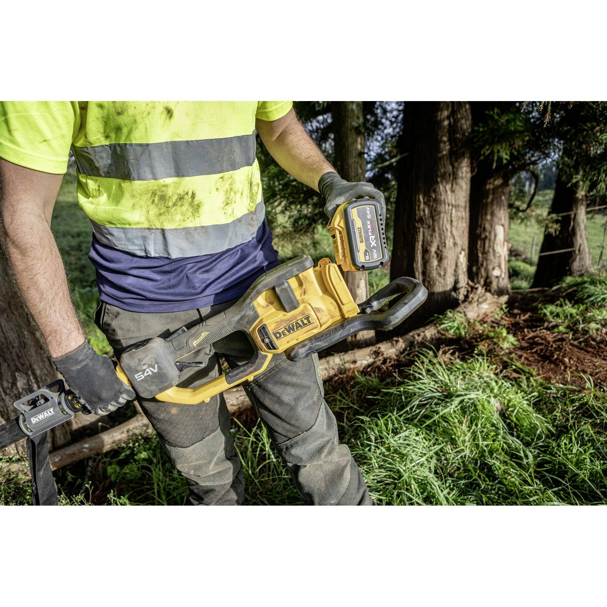 Mann in Arbeitskleidung hält eine Motorsäge im Wald. Er trägt Handschuhe und ein gelbes Sicherheitshemd. Bäume im Hintergrund.