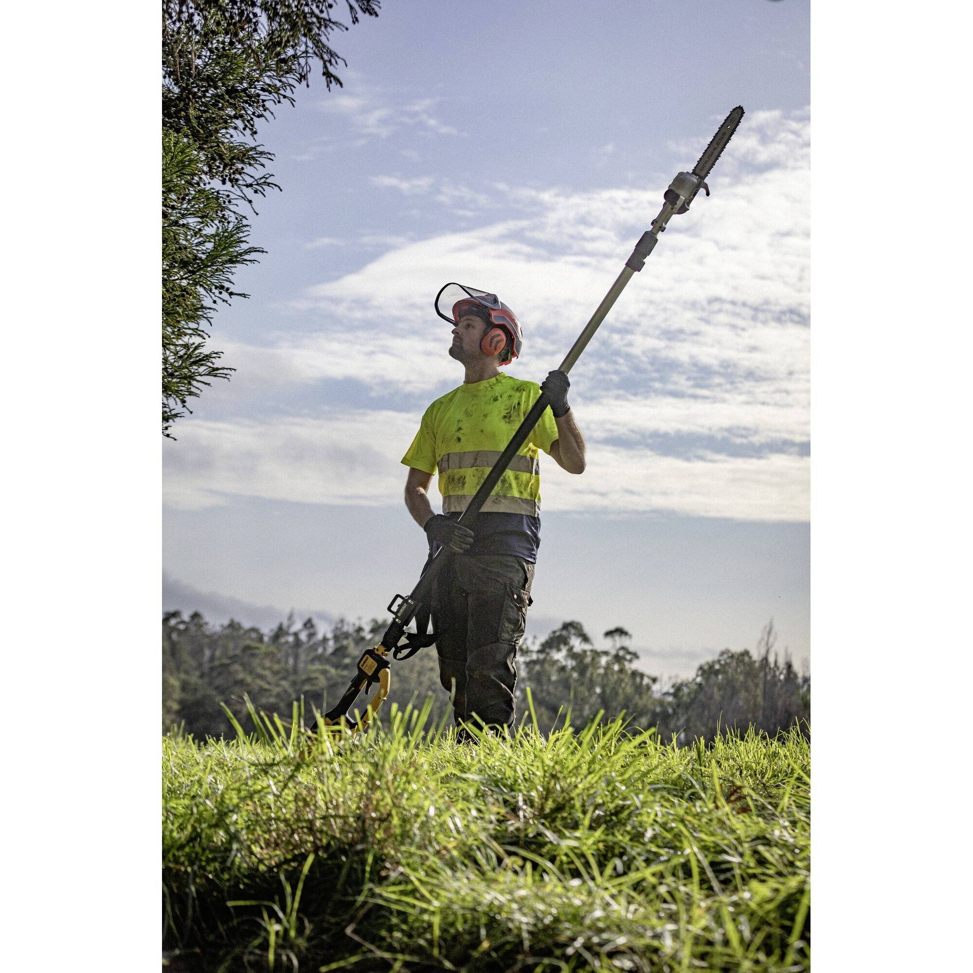 Der Mann trägt Schutzkleidung und hält eine Teleskop-Heckenschere. Er steht auf einer Wiese vor einem bewölkten Himmel.