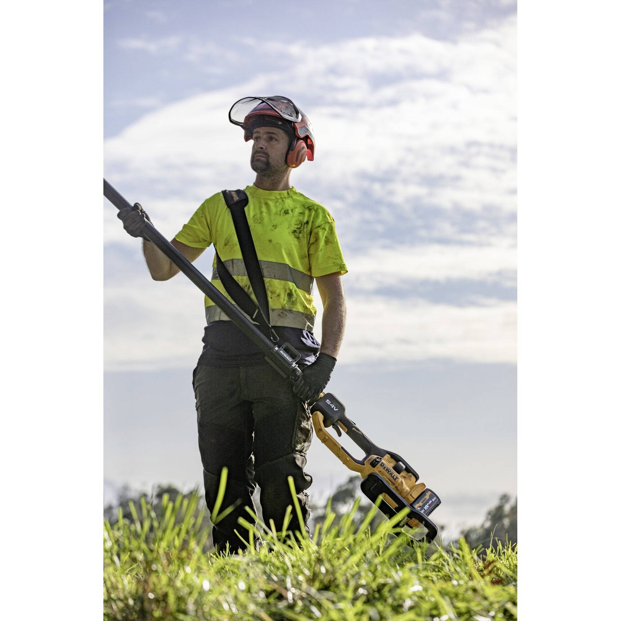 Eine Person im Sicherheitshelm und Warnweste schneidet mit einer Motorsense Gras auf einem Feld, helle Wolken am Himmel im Hintergrund.