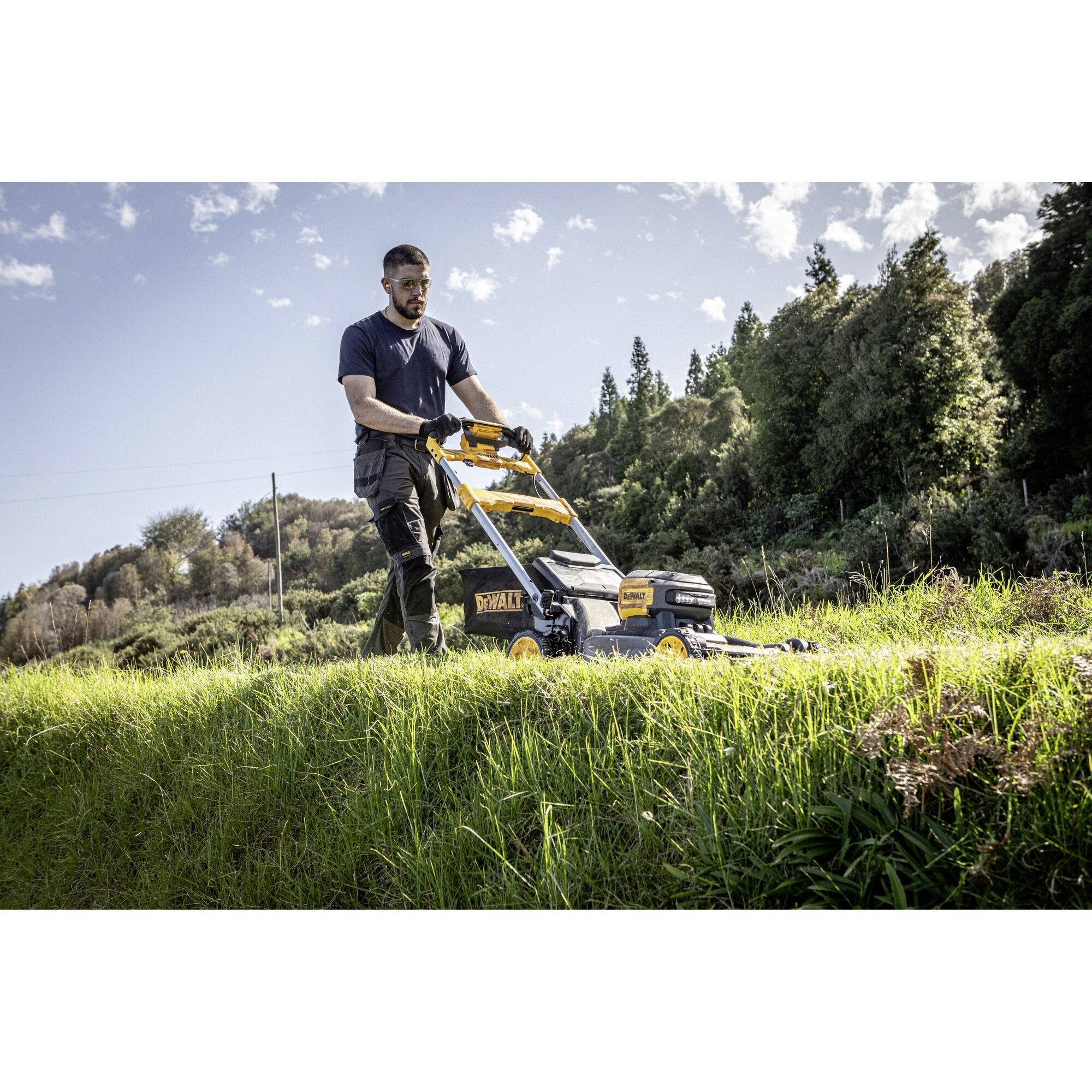Eine Person mäht mit einem gelben Rasenmäher auf einem grasbewachsenen Hang, umgeben von Bäumen und blauem Himmel.