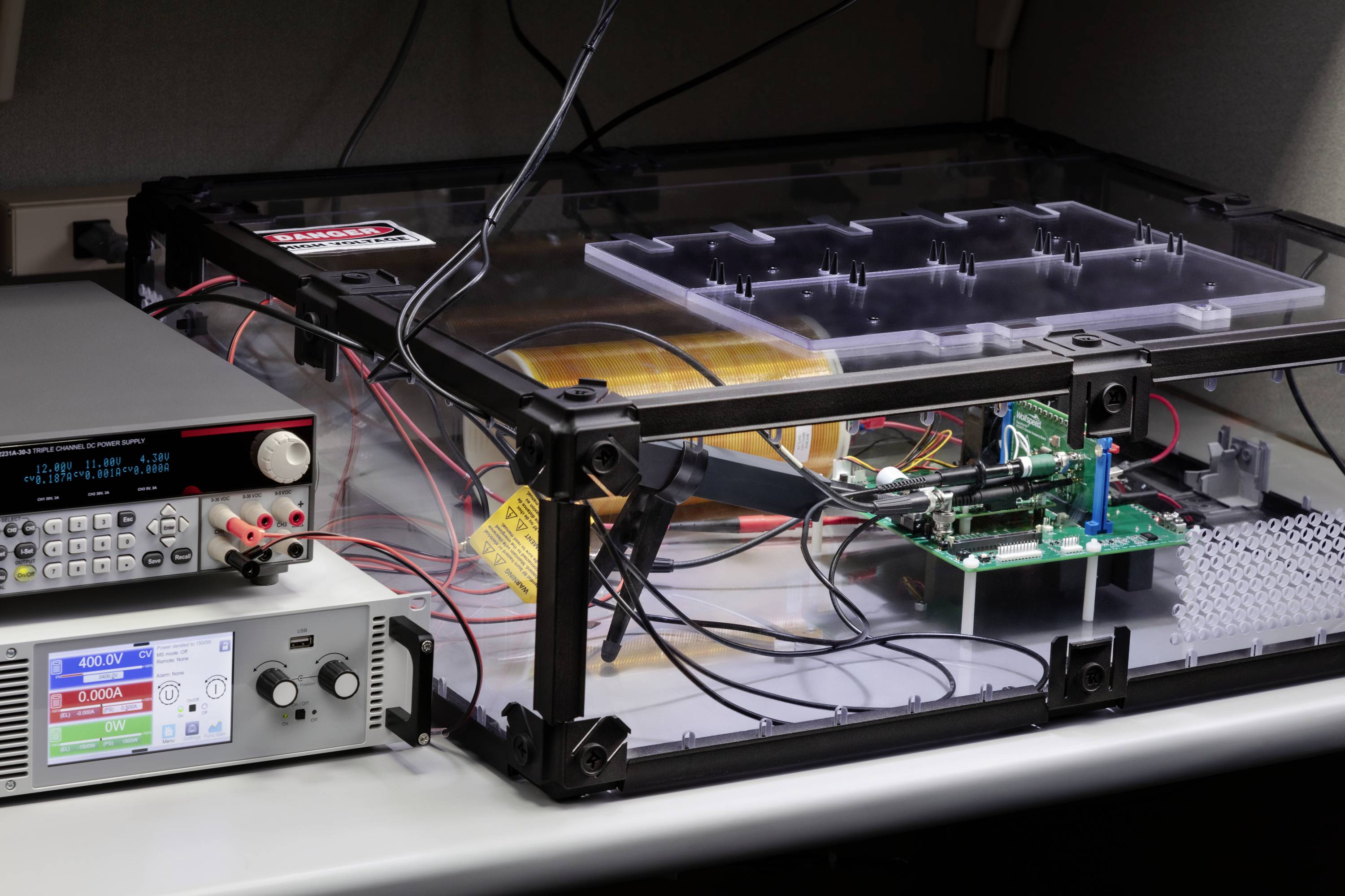 Elektroniktestaufbau auf einem Tisch mit Geräten und Leiterplatten in einem durchsichtigen Gehäuse, umgeben von Kabeln und Anzeigen.