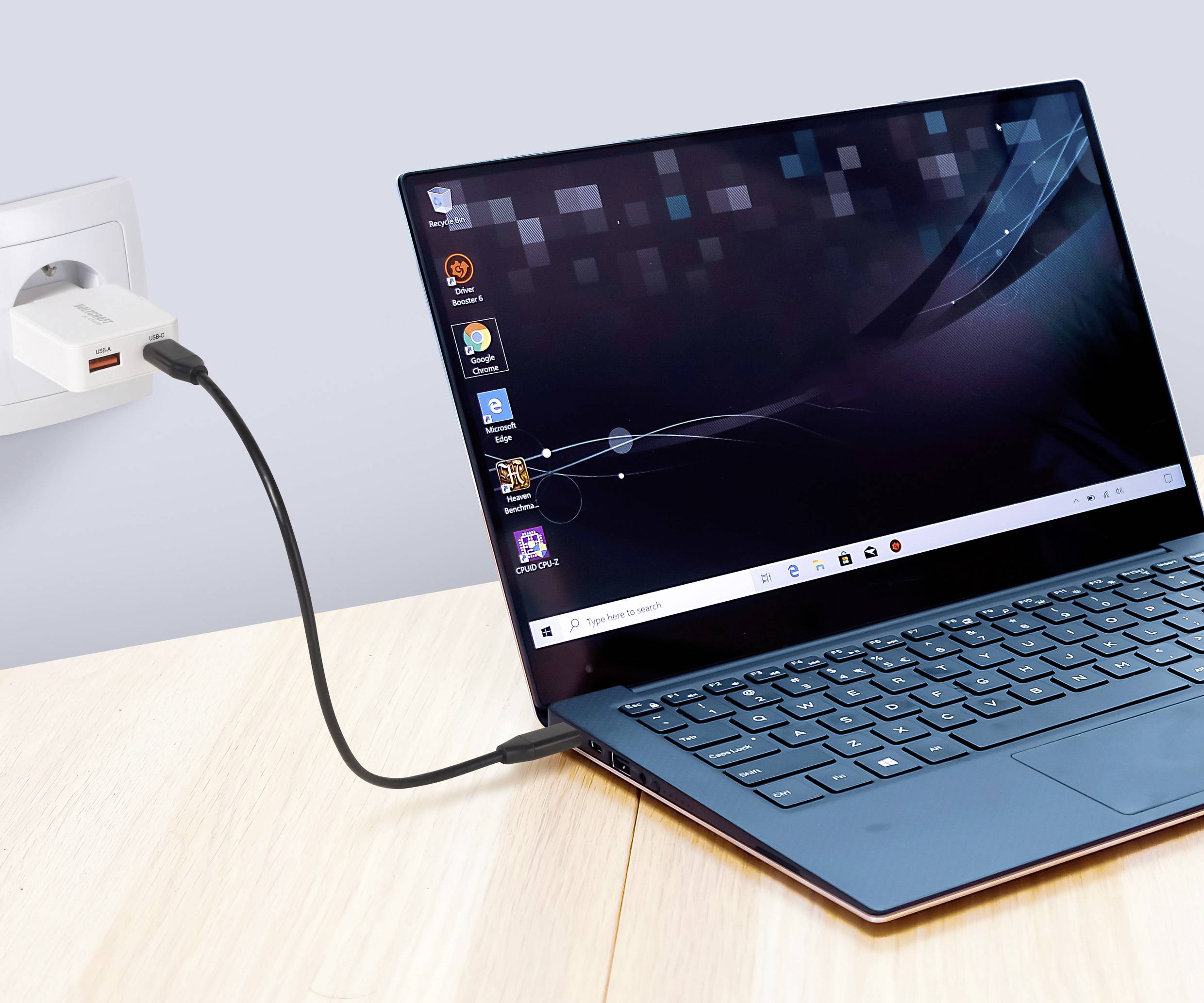 Ein blauer Laptop, der auf einem Holztisch lädt und an eine Wandsteckdose angeschlossen ist. Der Bildschirm zeigt verschiedene Anwendungen vor einem Desktop-Hintergrund.