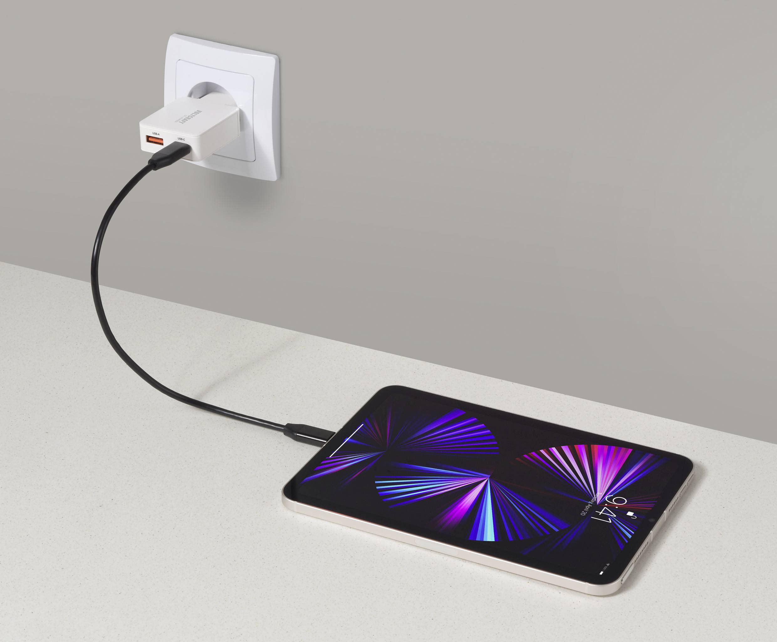 Ein Tablet, das auf einer Arbeitsfläche lädt und über ein USB-C-Kabel und Ladegerät mit einer Wandsteckdose verbunden ist. Das Tablet zeigt ein farbenfrohes abstraktes Design.