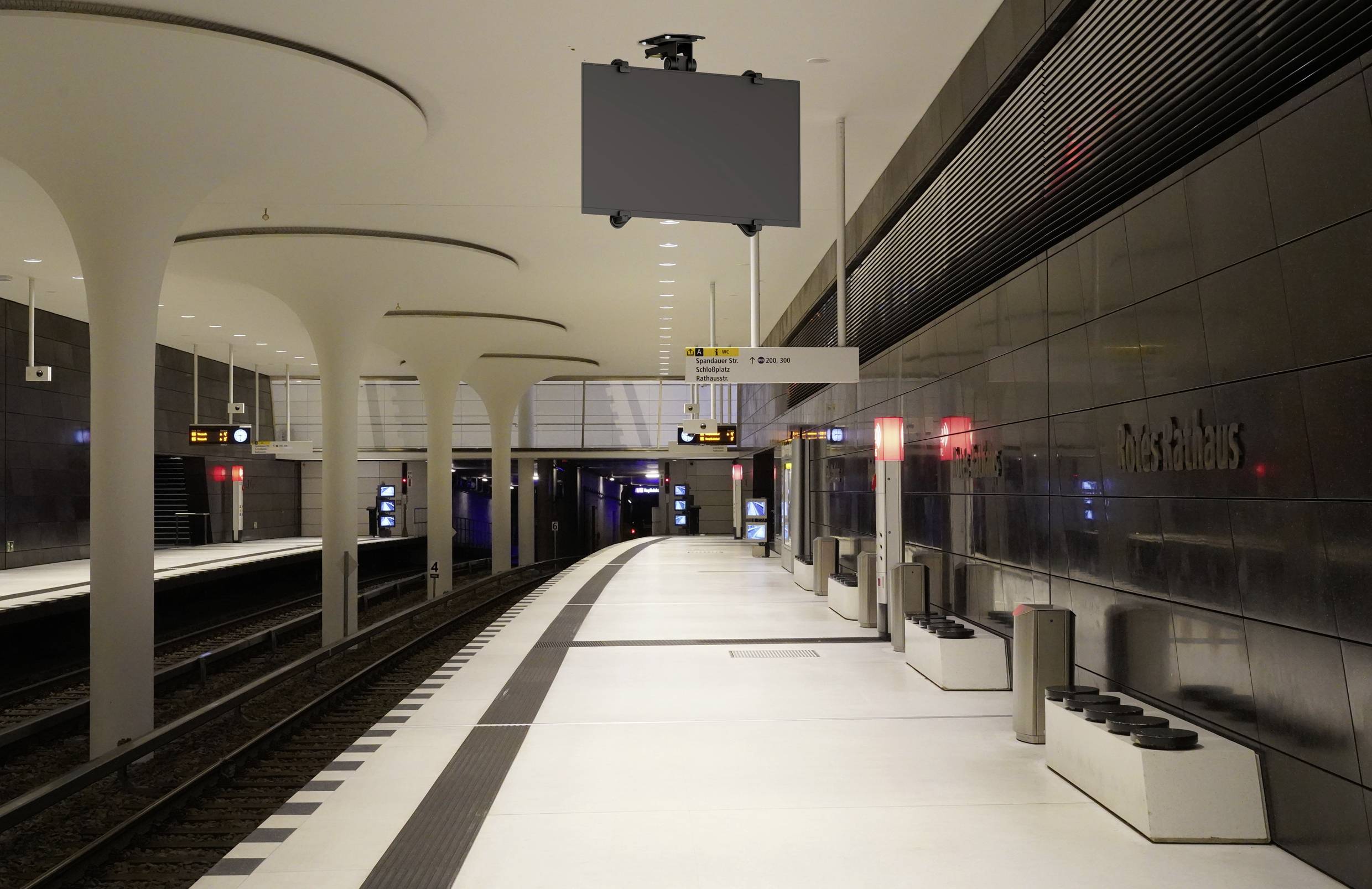 Leerer moderner U-Bahnhof mit weißen Böden, hohen Säulen und dunklen Wänden. Gleise auf der linken Seite, keine Personen sichtbar.
