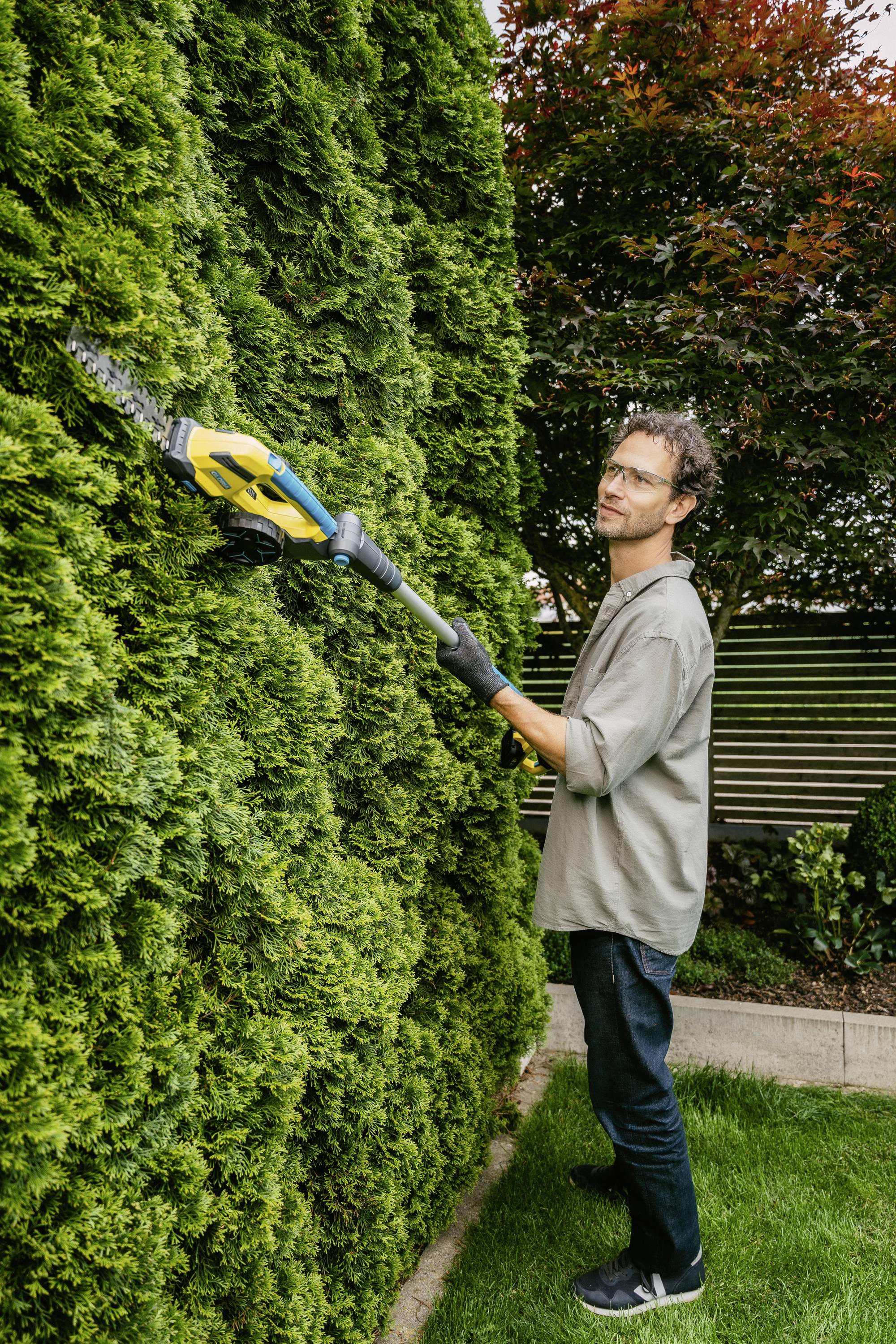 Eine Person trimmt eine grüne Hecke mit einer elektrischen Heckenschere im Garten, umgeben von üppiger Vegetation und einem Holzzaun.