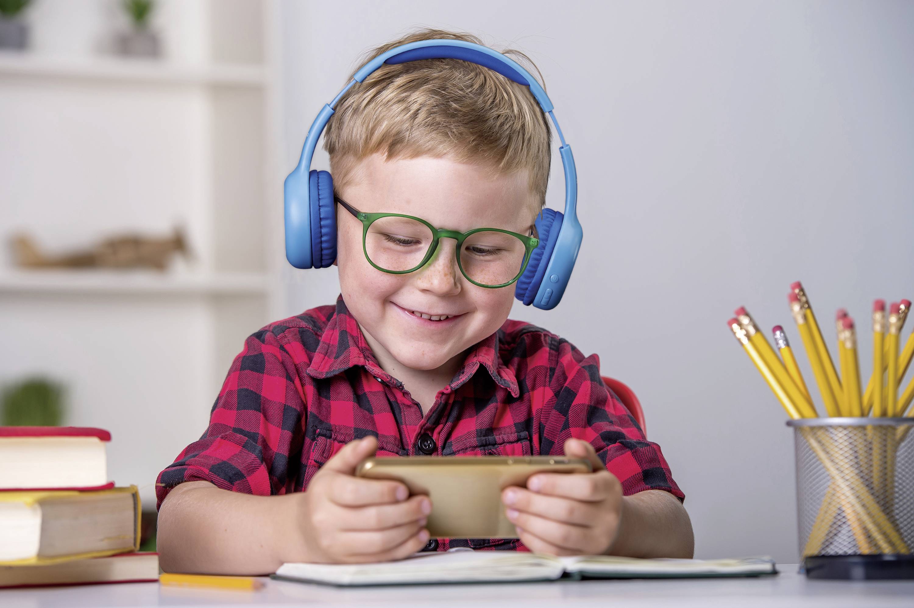 Ein lächelnder Junge mit Brille und blauen Kopfhörern spielt an einem Smartphone am Tisch. Neben ihm liegen Bücher und ein Stiftehalter.