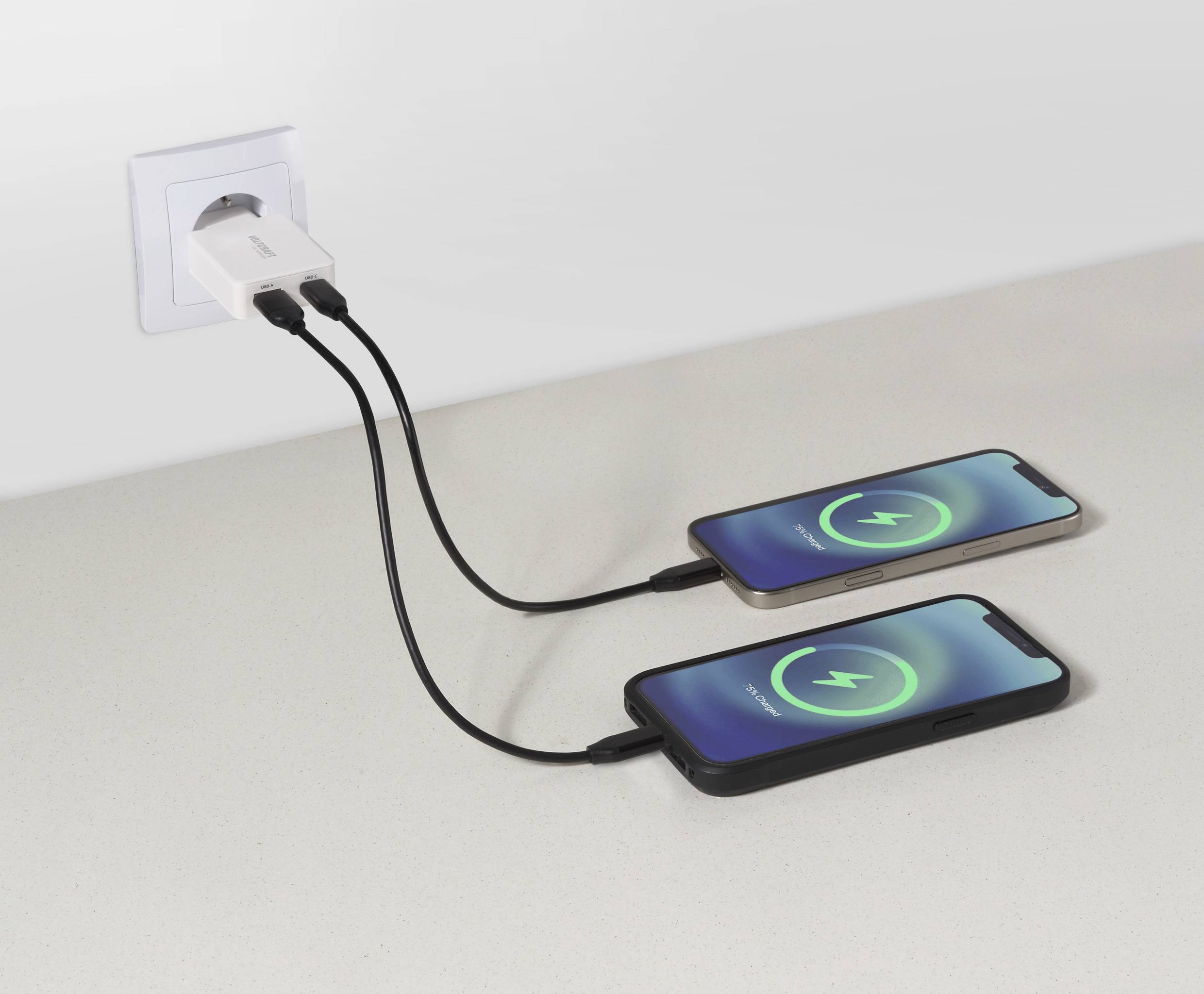 Zwei Smartphones auf einem Tisch, die an einem Wandadapter angeschlossen sind, beide mit einem grünen Ladezeichen auf ihren Bildschirmen, was eine aktive Aufladung anzeigt.