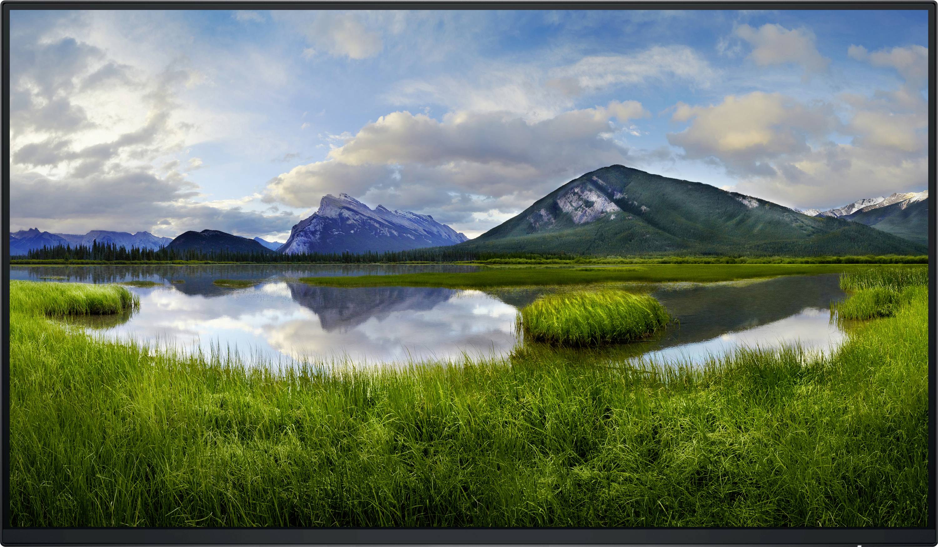 Panoramablick auf Berglandschaft mit stillem See im Vordergrund, umgeben von grünen Wiesen und wolkigem Himmel.