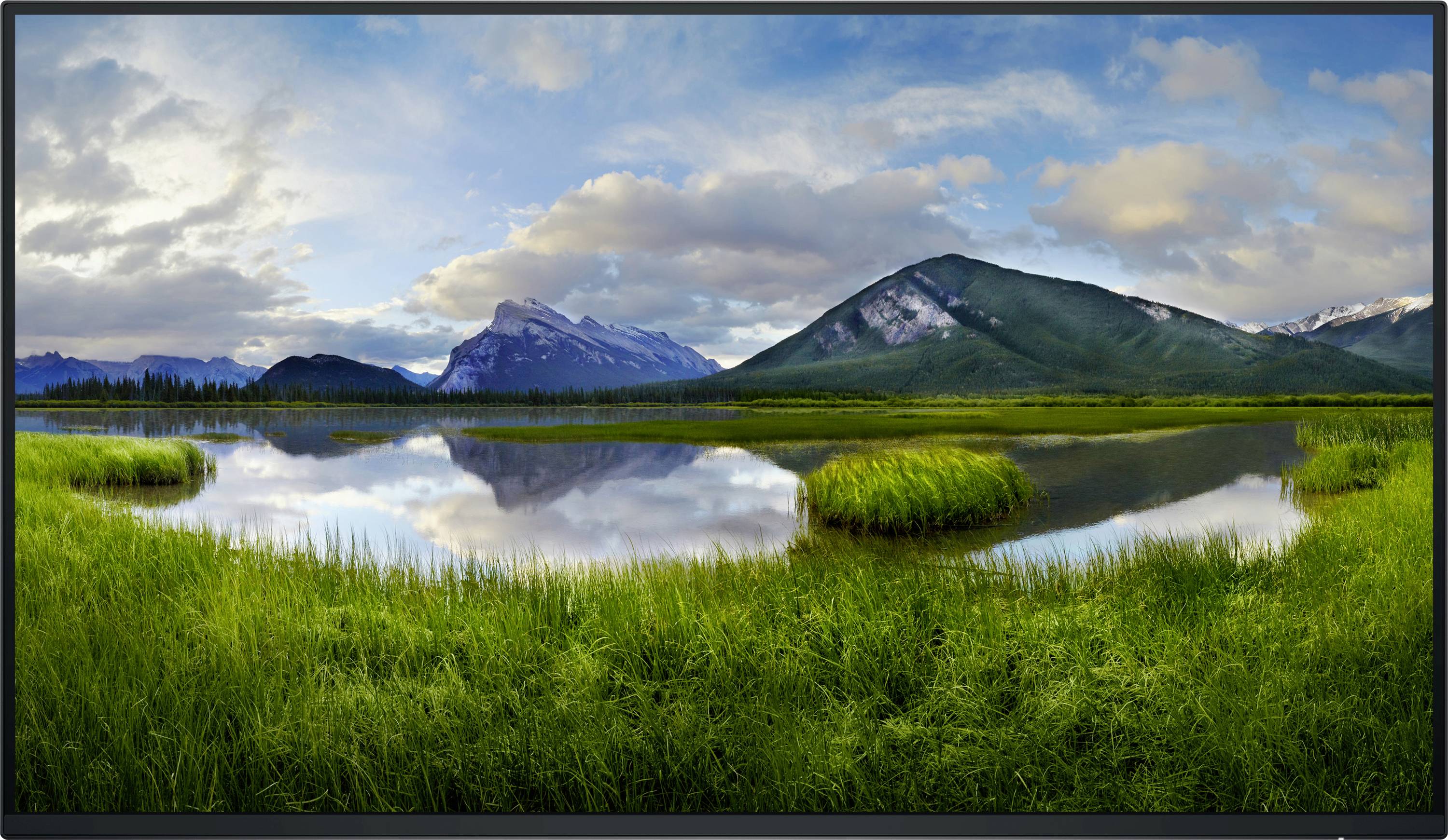 Berglandschaft mit einem See im Vordergrund, umgeben von Gras und Bergen im Hintergrund unter bewölktem Himmel.
