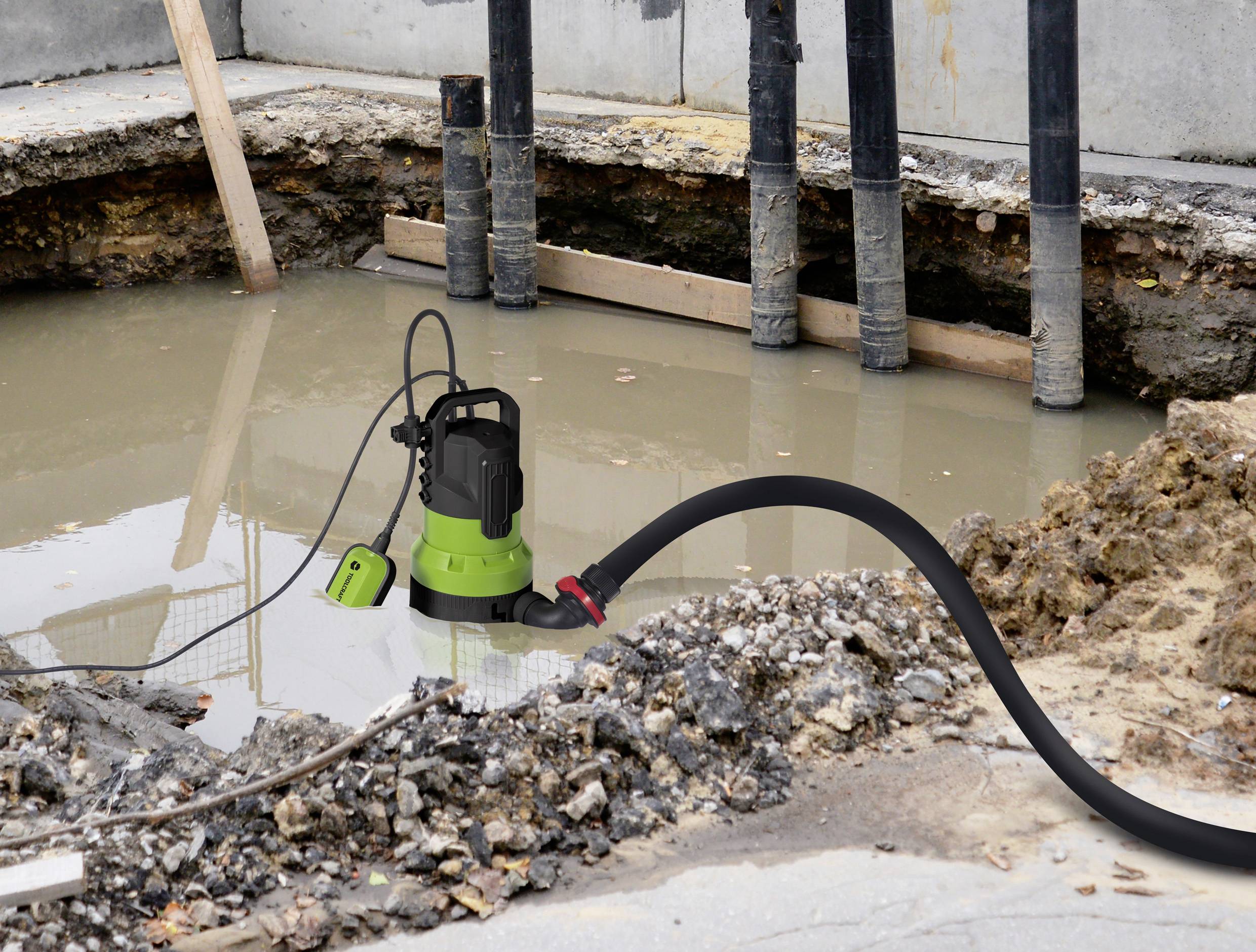 Eine grüne Tauchpumpe entwässert eine Baustelle mit schlammigem Wasser. Schwarzer Schlauch führt von der Pumpe ab.