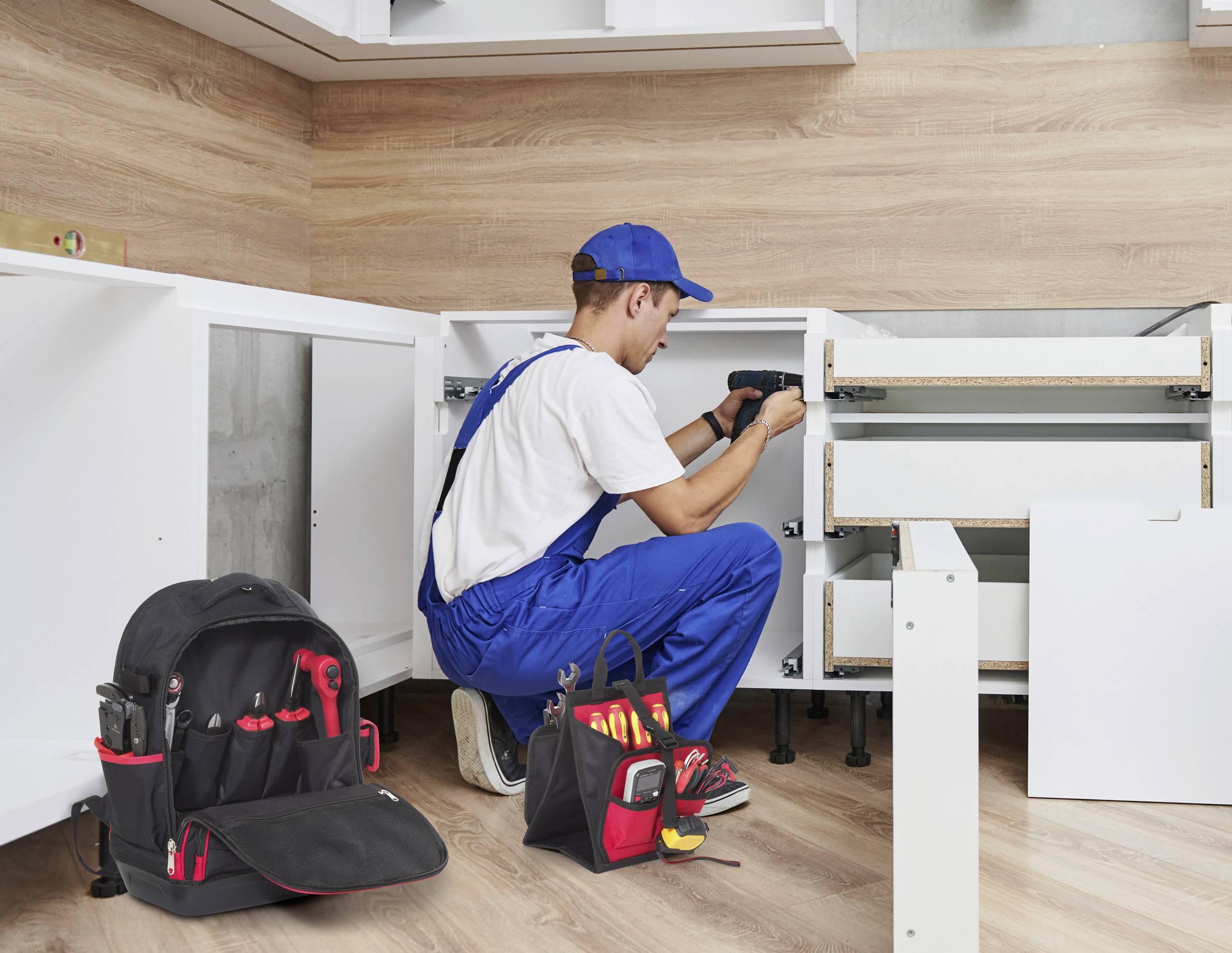 Ein Handwerker in blauen Overalls und einer Kappe montiert Küchenschränke mit einer Akkuschrauber. Eine Werkzeugtasche mit roten und gelben Werkzeugen liegt in der Nähe.