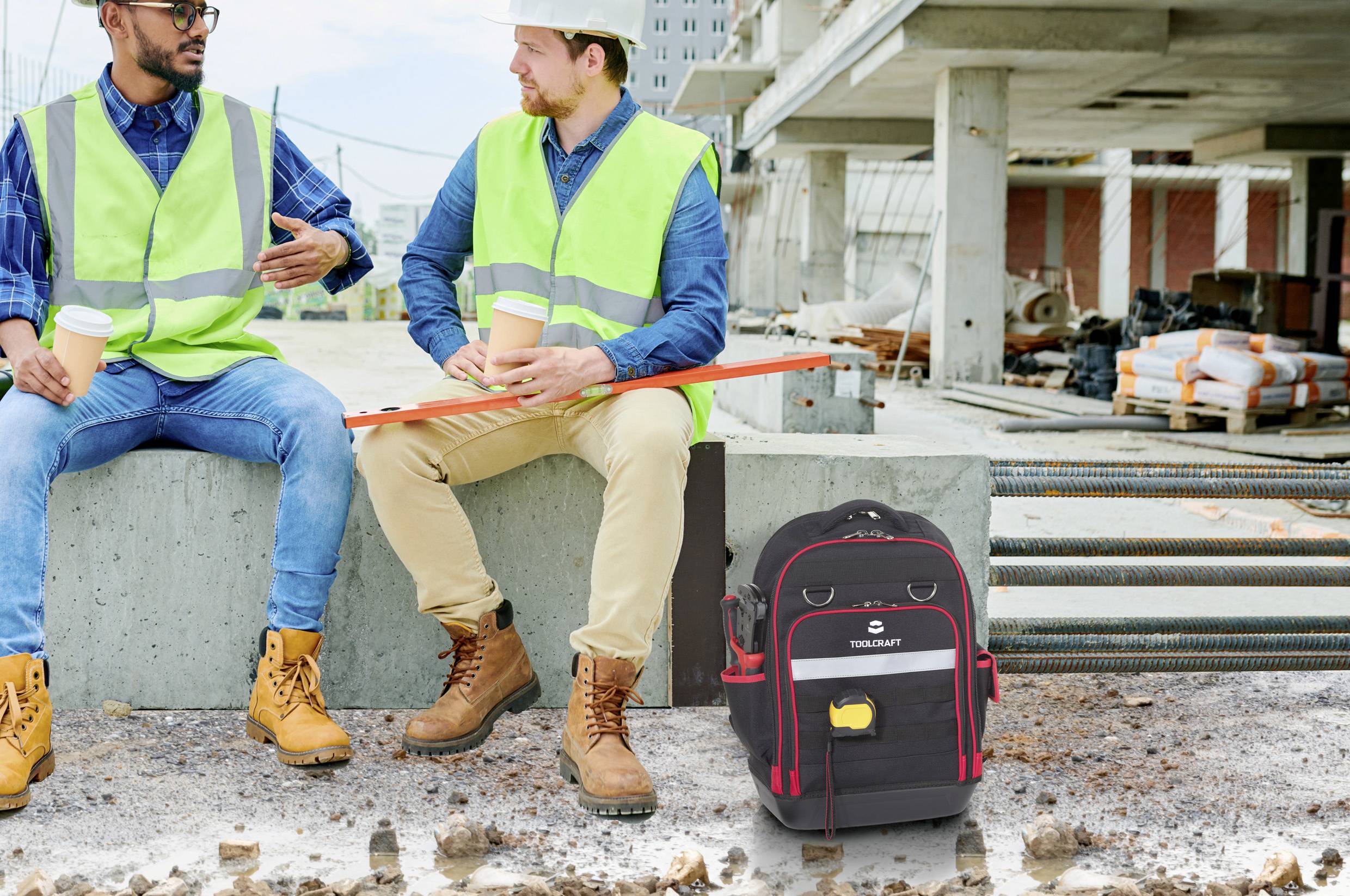 Zwei Bauarbeiter in Sicherheitswesten und Helmen sitzen auf einem Betonblock auf einer Baustelle und unterhalten sich. Ein Rucksack liegt auf dem Boden.