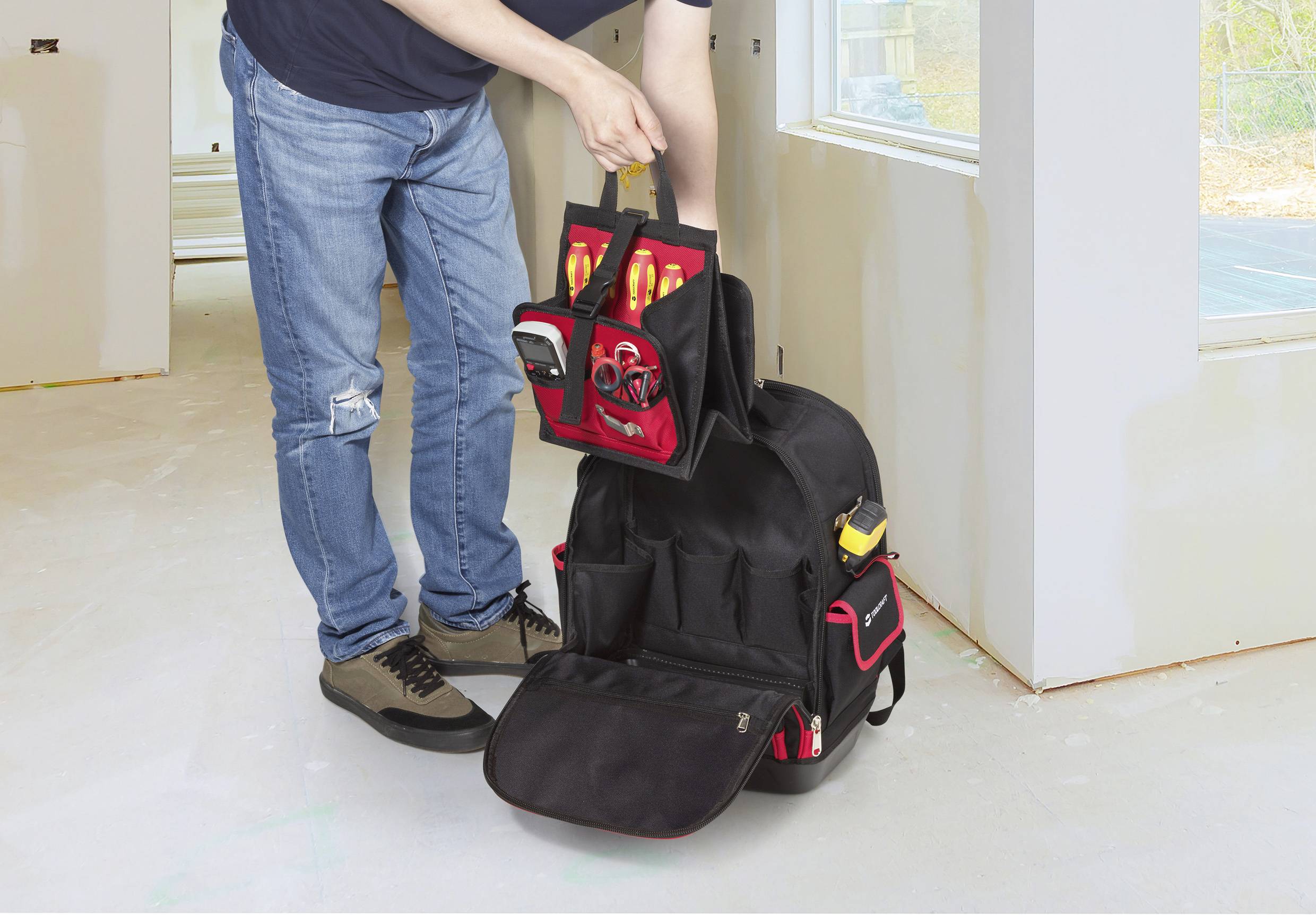 Eine Person, die Werkzeuge in einem schwarzen Rucksack mit roten Akzenten ordnet, in einem gut beleuchteten Raum im Rohbau.