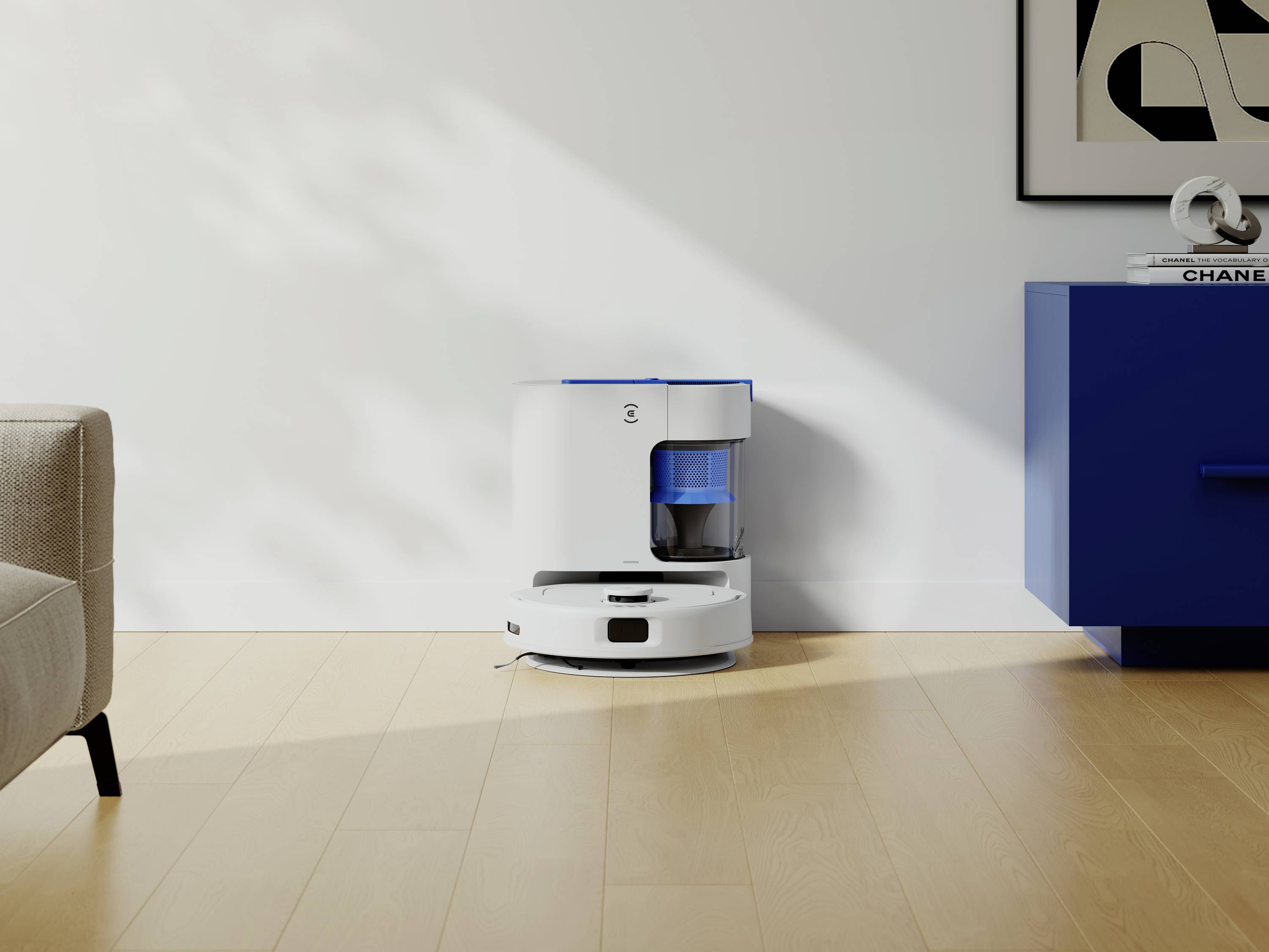 Ein weißer Saugroboter auf einem Holzboden, neben einer blauen Kommode und einem Sofa. An der Wand hängt ein abstraktes Kunstwerk.