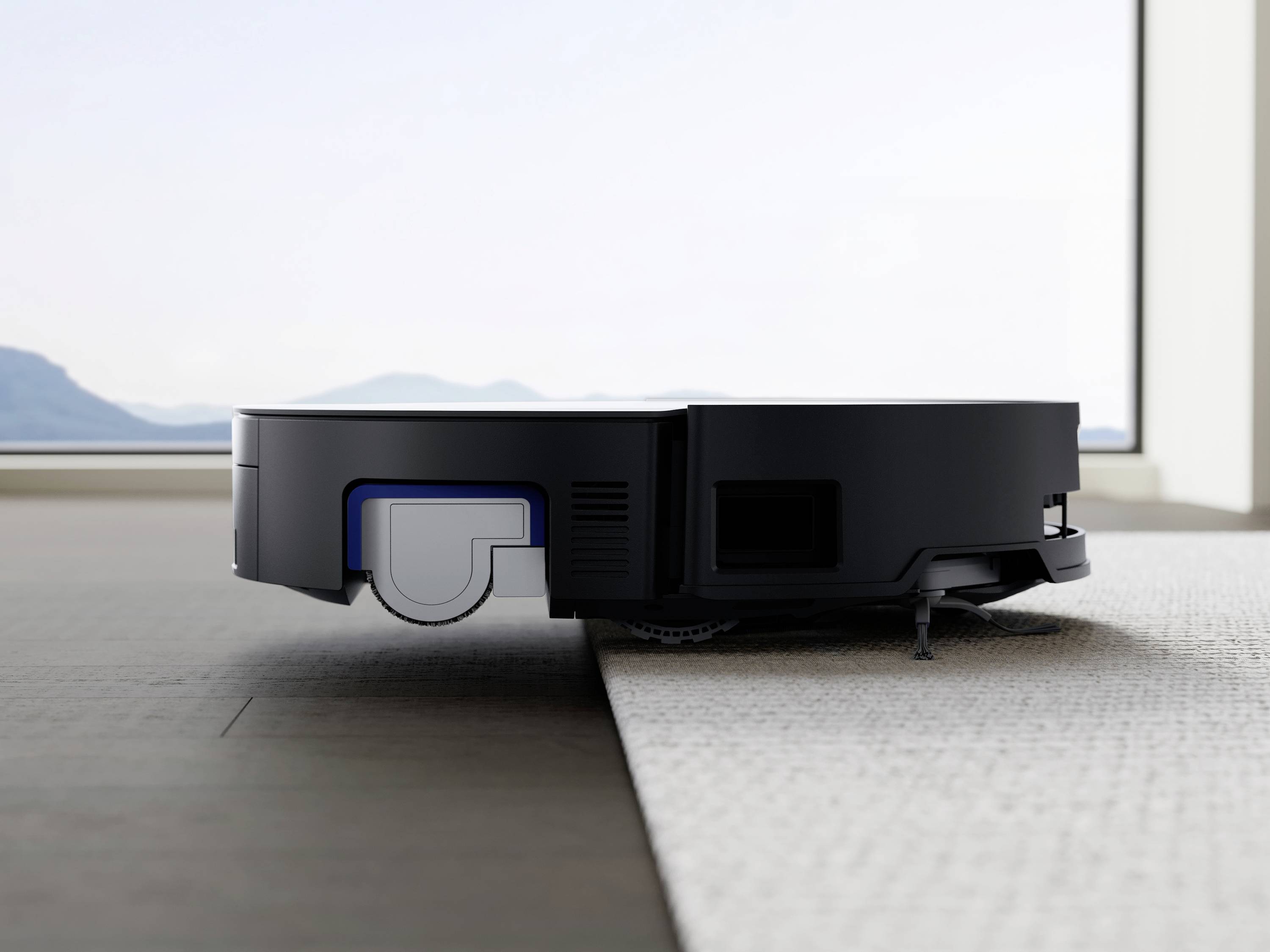 Ein Saugroboter bewegt sich von einem harten Holzboden auf einen Teppich in einem modern eingerichteten Raum mit Aussicht auf Berge.