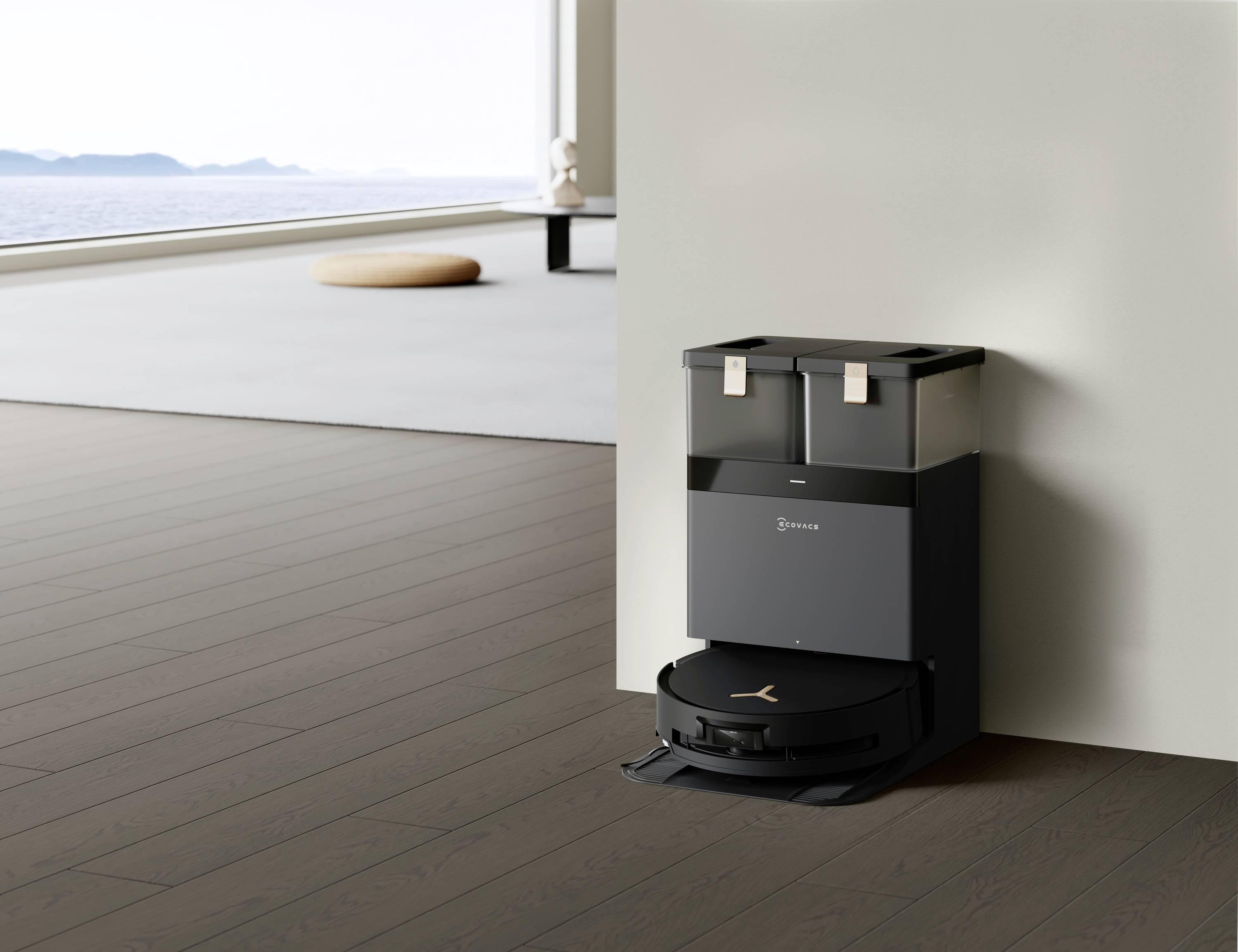 Ein schwarzer Saugroboter in einer modernen Wohnungsecke ladet an einer Station. Große Fenster bieten Blick auf das Meer.