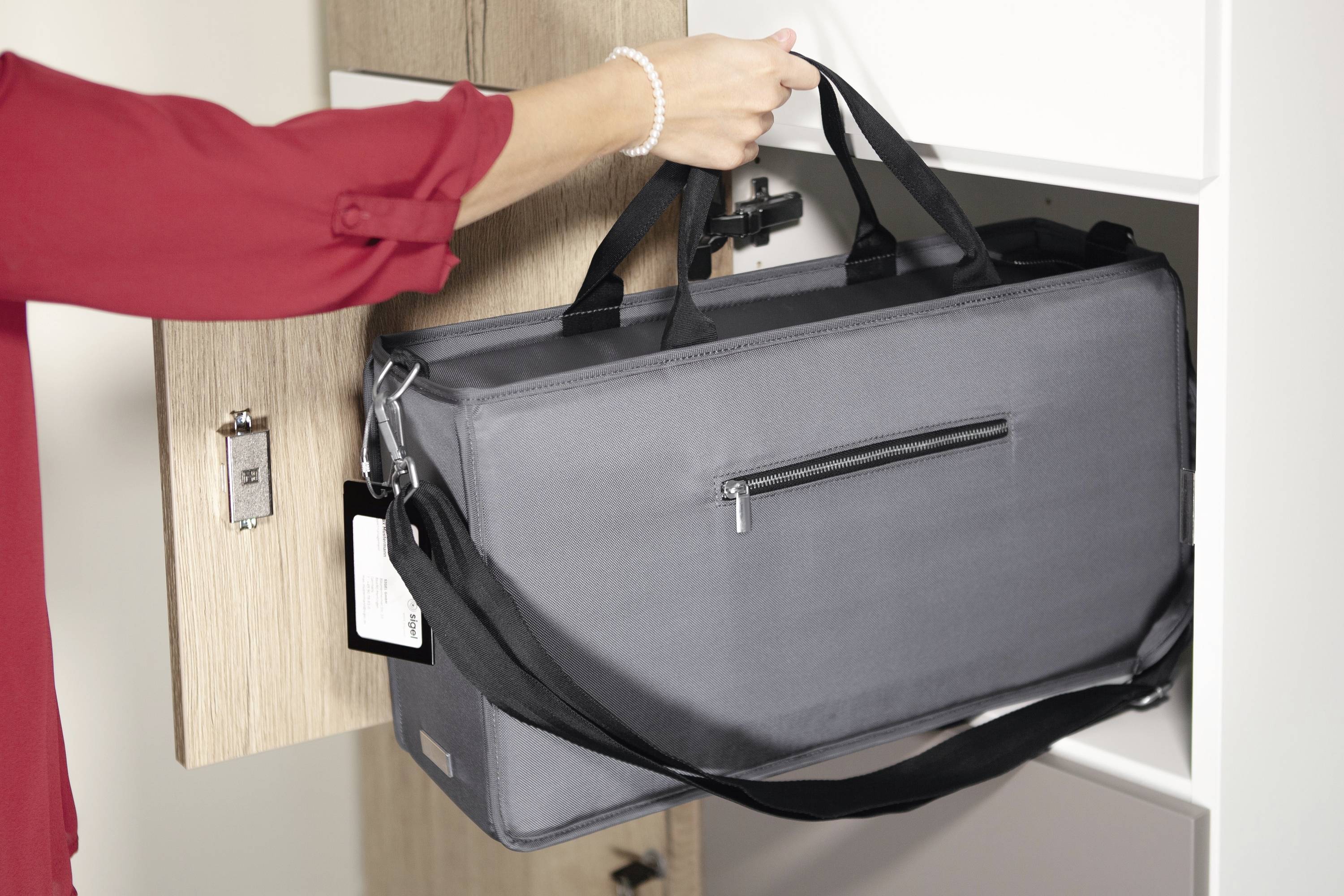 Eine Person in roter Bluse verstaut eine graue Tragetasche in einem Schrank. Die Tasche hat Tragegriffe und einen Reißverschluss.