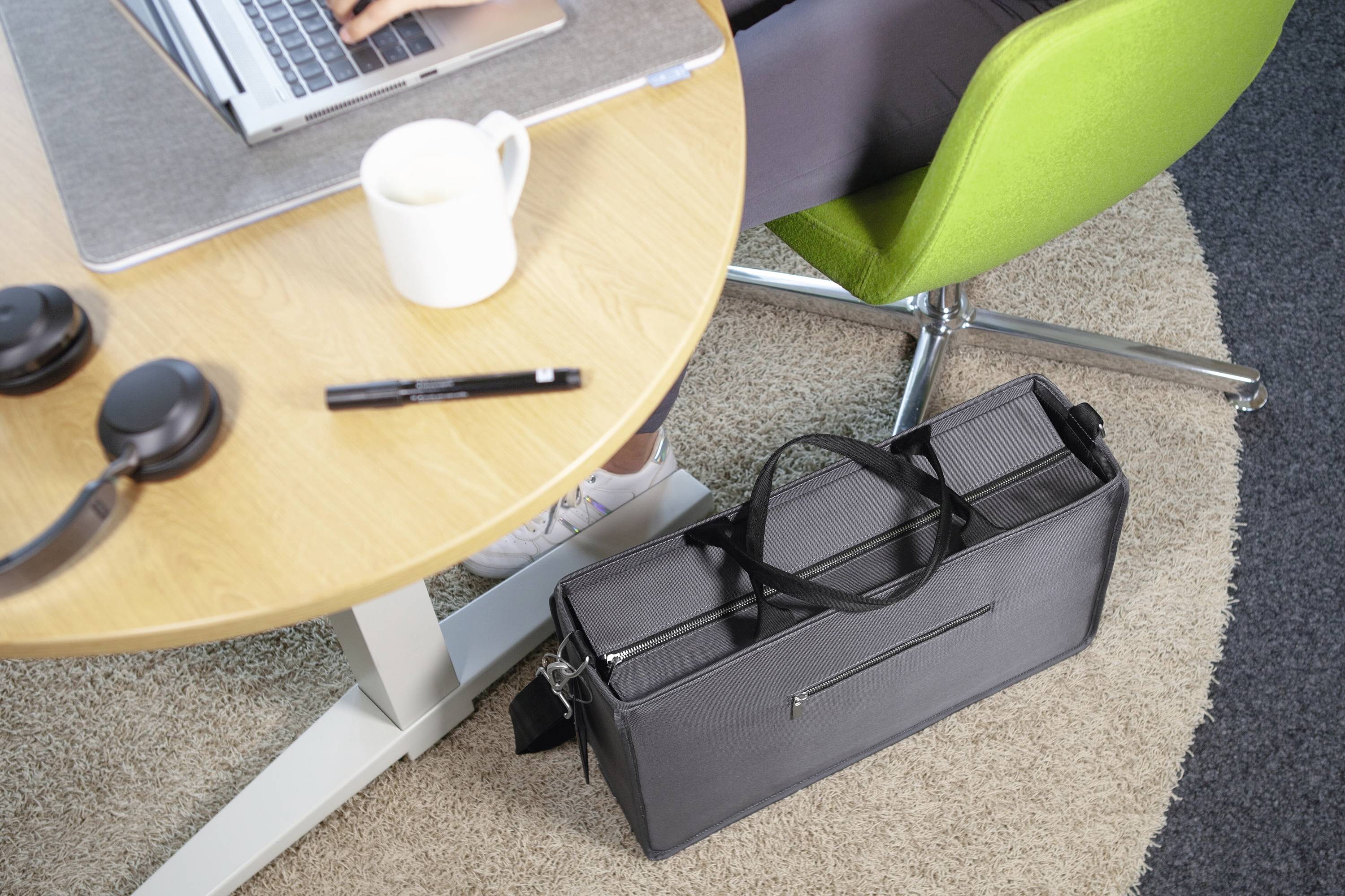 Eine Person arbeitet an einem Laptop an einem runden Tisch. Auf dem Tisch stehen eine Tasse, Kopfhörer und ein Stift. Daneben steht eine schwarze Tasche.